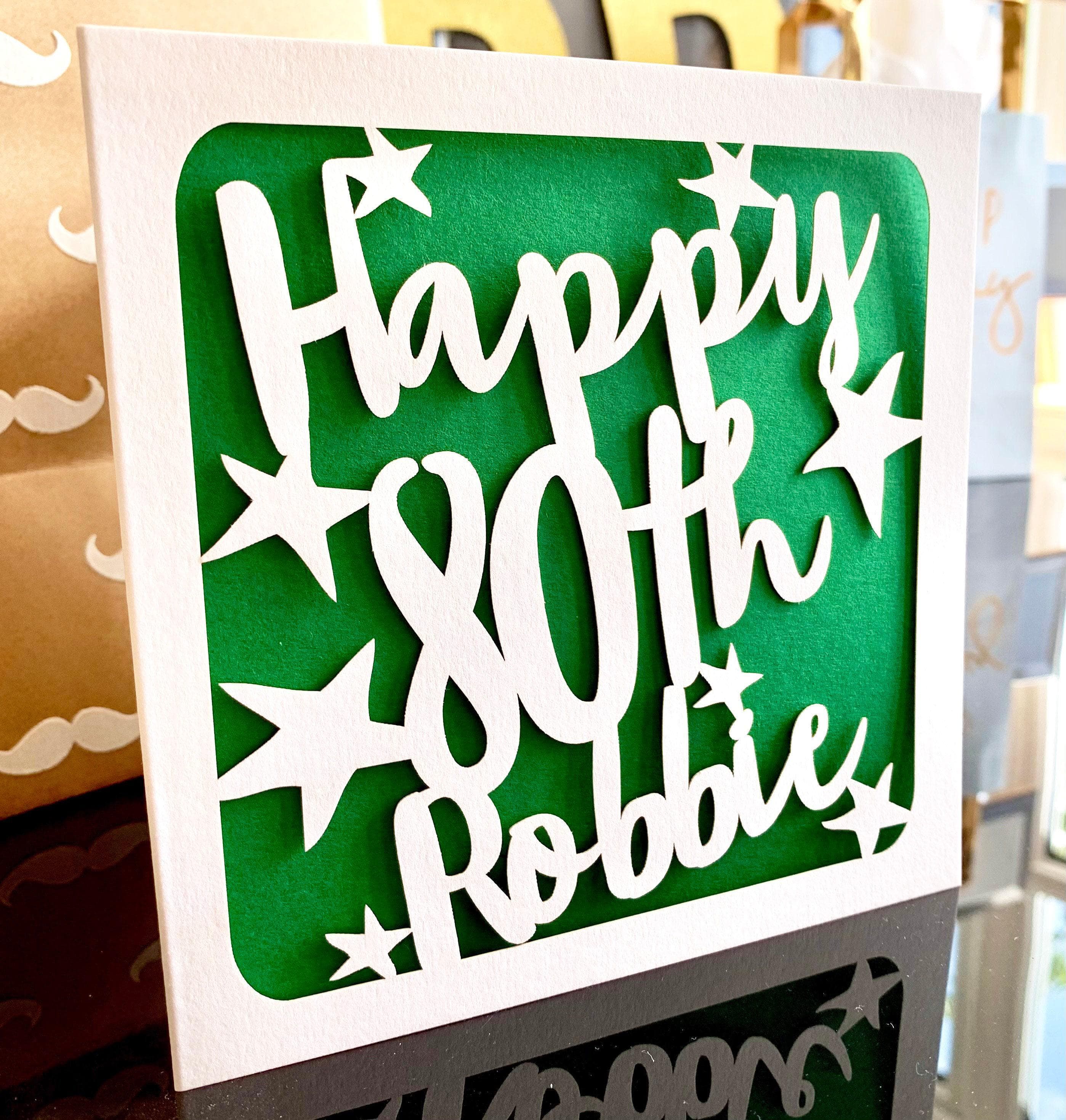 White square laser cut 80th birthday card. Featuring the words ‘Happy 80th’ cut in a bold script font across the card, which has been personalised with a name in the last line. Hand-drawn stars surround the text. The card has a dark green paper insert and is standing on a shiny black surface, with gifts visible behind.