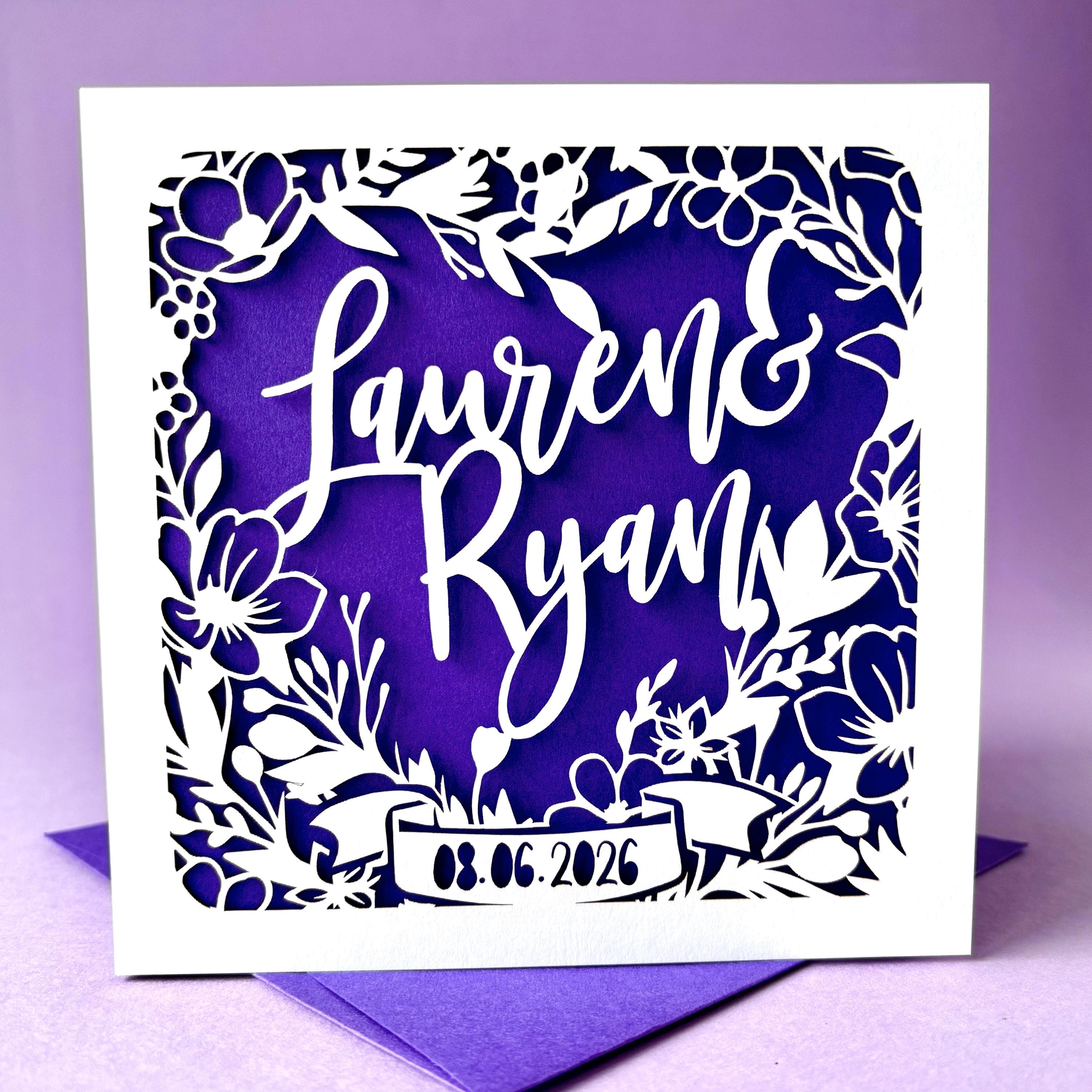 Square white laser cut wedding card featuring a heart shaped floral wreath. The centre of the wreath is personalised with two names in an elegant script font. The base of the wreath features a ribbon banner, with a date showing across it. The card has a purple paper insert, and is standing on a colour-matched envelope. The card is standing against a pale purple background.