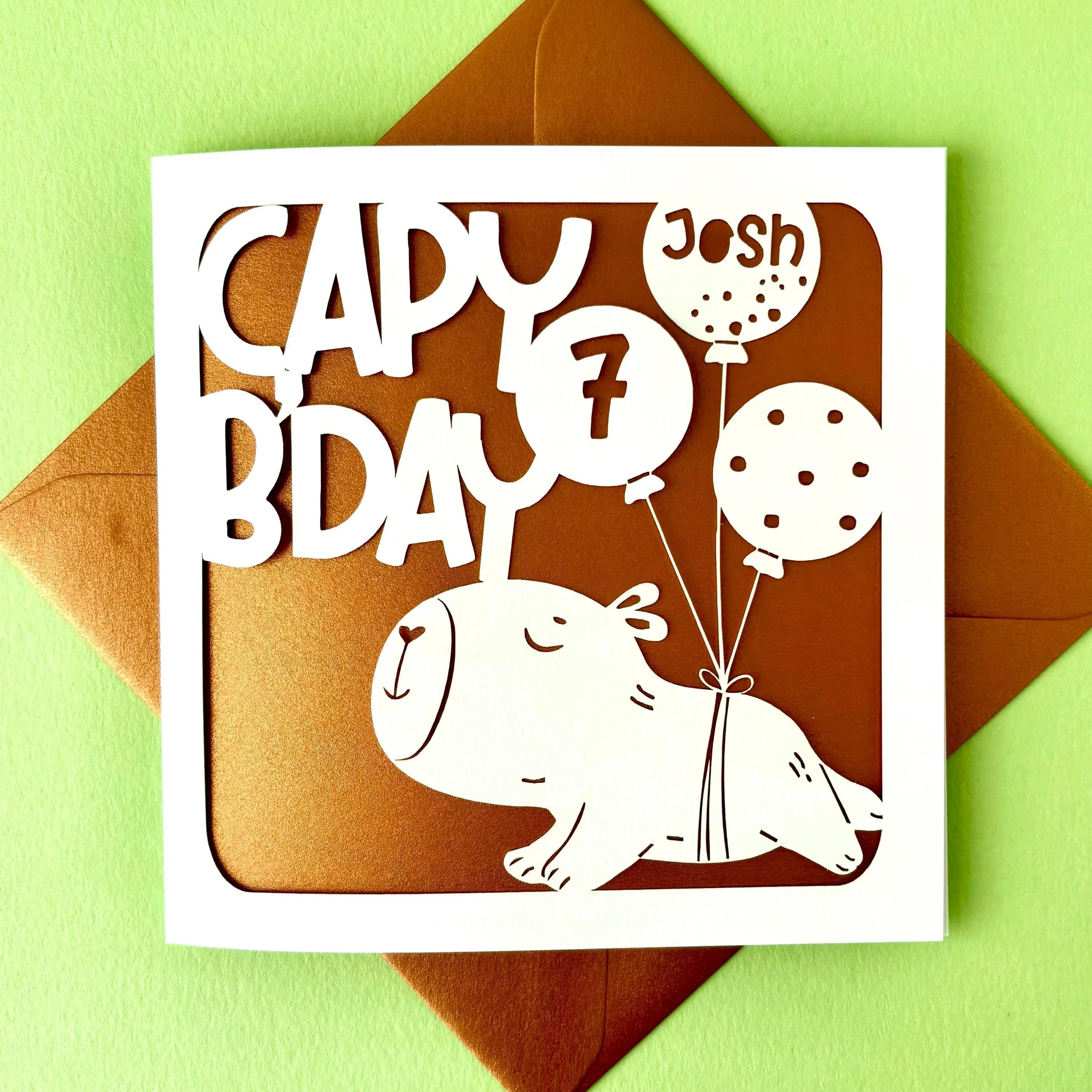 White square laser cut kids birthday card. Featuring a cute cartoon capybara cut into the card, being lifted by three balloons. One balloon has a spots pattern, another has been personalised with a name, and the third with an age. The top of the card reads 'CAPY B'DAY' in a bold playful font. The card has a bronze paper insert and is lying on a colour-matched envelope. The background is pale green.