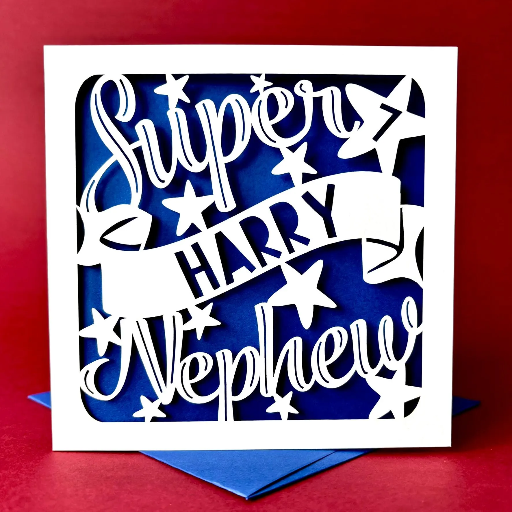 White square laser cut Nephew birthday card. Featuring the words 'Super Nephew' in a bold script font across the top and bottom of the card, personalised with a name displayed on a banner in the centre. Hand‑drawn stars surround the design. The card has a dark blue paper insert and is standing on a colour-matched envelope against a deep red background.