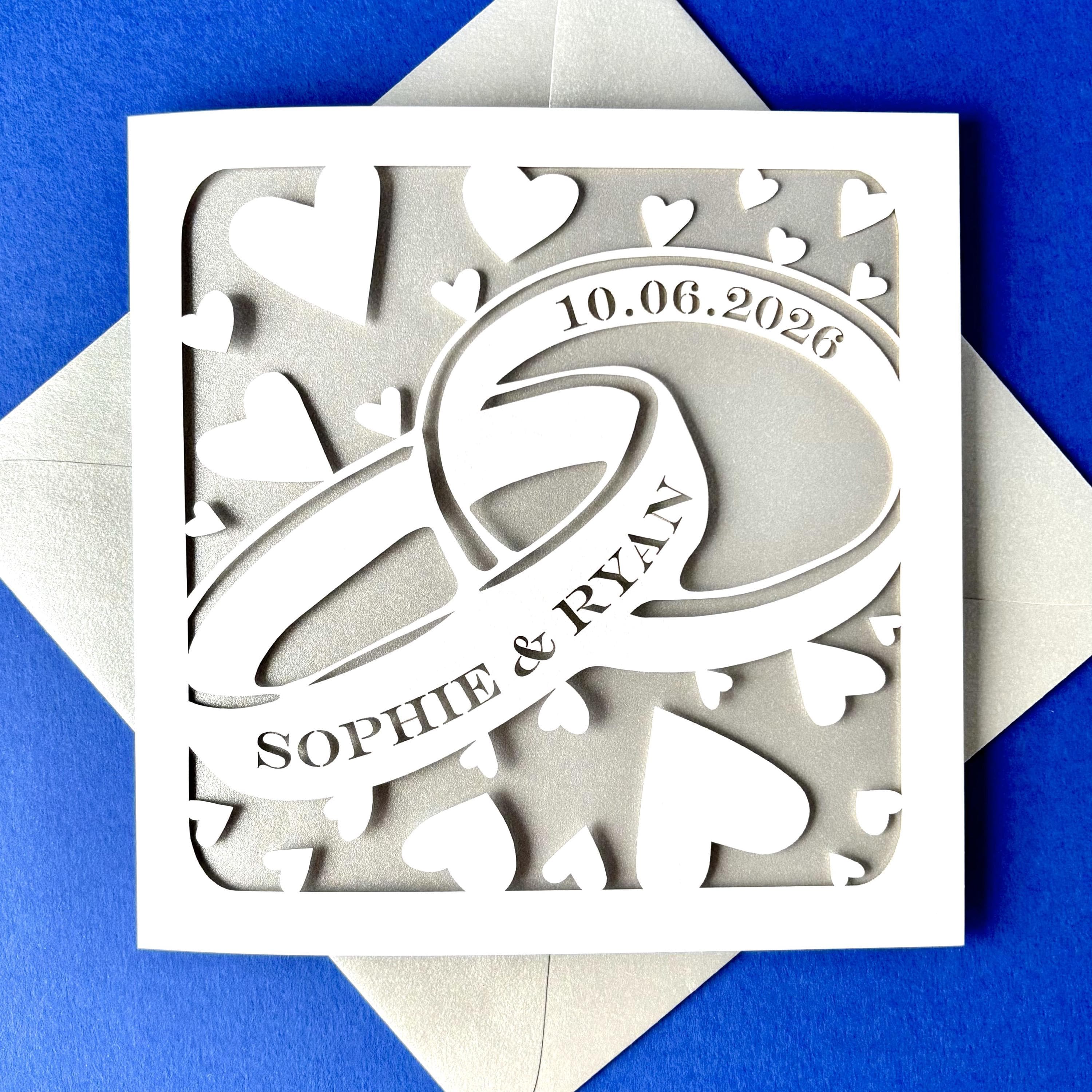 Square white laser cut greetings card featuring two entwined rings surrounded by hearts. The front ring has been personalised with two names, and the second with a date, in a traditional serif font. The wedding card has a silver paper insert and is lying on a matching envelope. It is lying on a bright blue background.