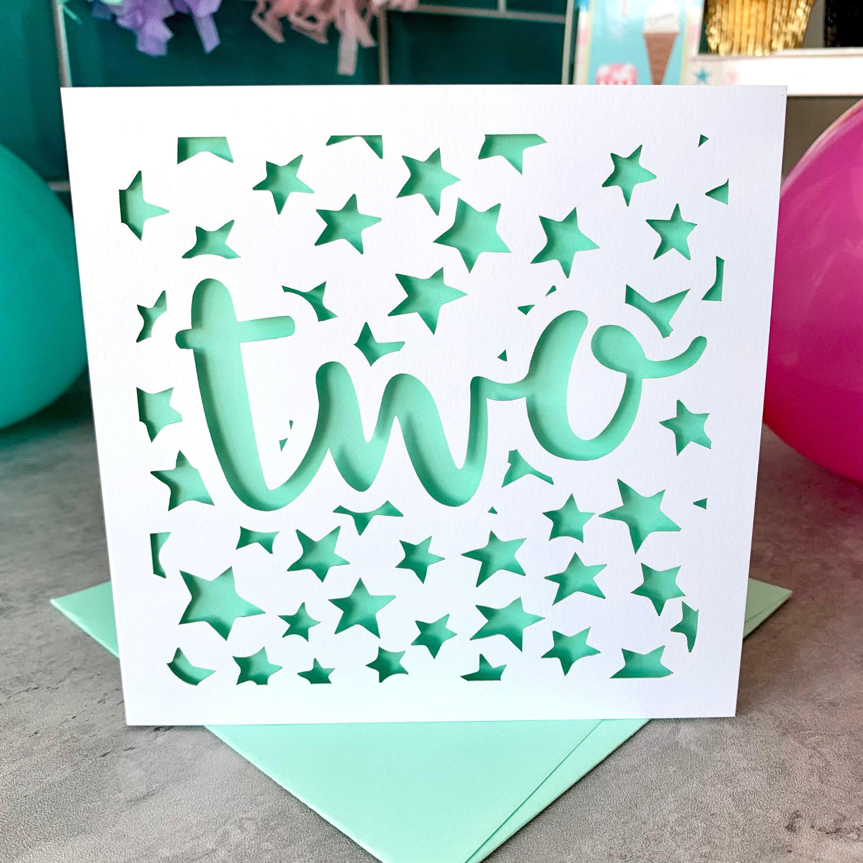 White square laser cut 2nd birthday card. Featuring a stars pattern cut across the background, with the large word two cut across the centre in a bold script font. The card has a mint paper insert and is standing on a colour-matched envelope. The card is standing against a festive background, with cupcakes and balloons visible behind.