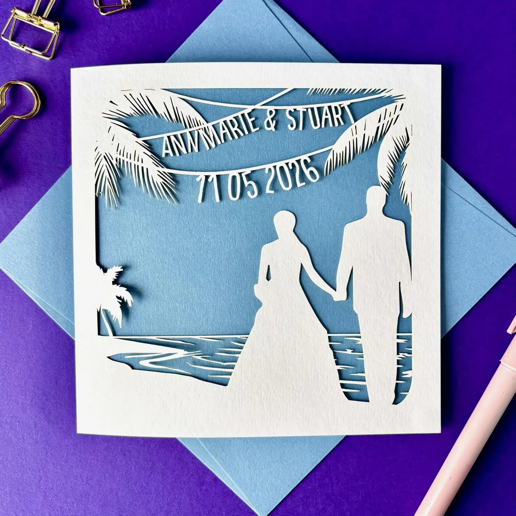 White square laser cut wedding card featuring the silhouette of a bride and groom wedding couple on a beach, surrounded by palm trees. Above the couple the card has been personalised with bunting which shows their name and the date of the wedding. The card has a pale blue paper insert and is lying flat on a colour matched envelope. It is lying against a purple background, with stationery items visible surorunding it.