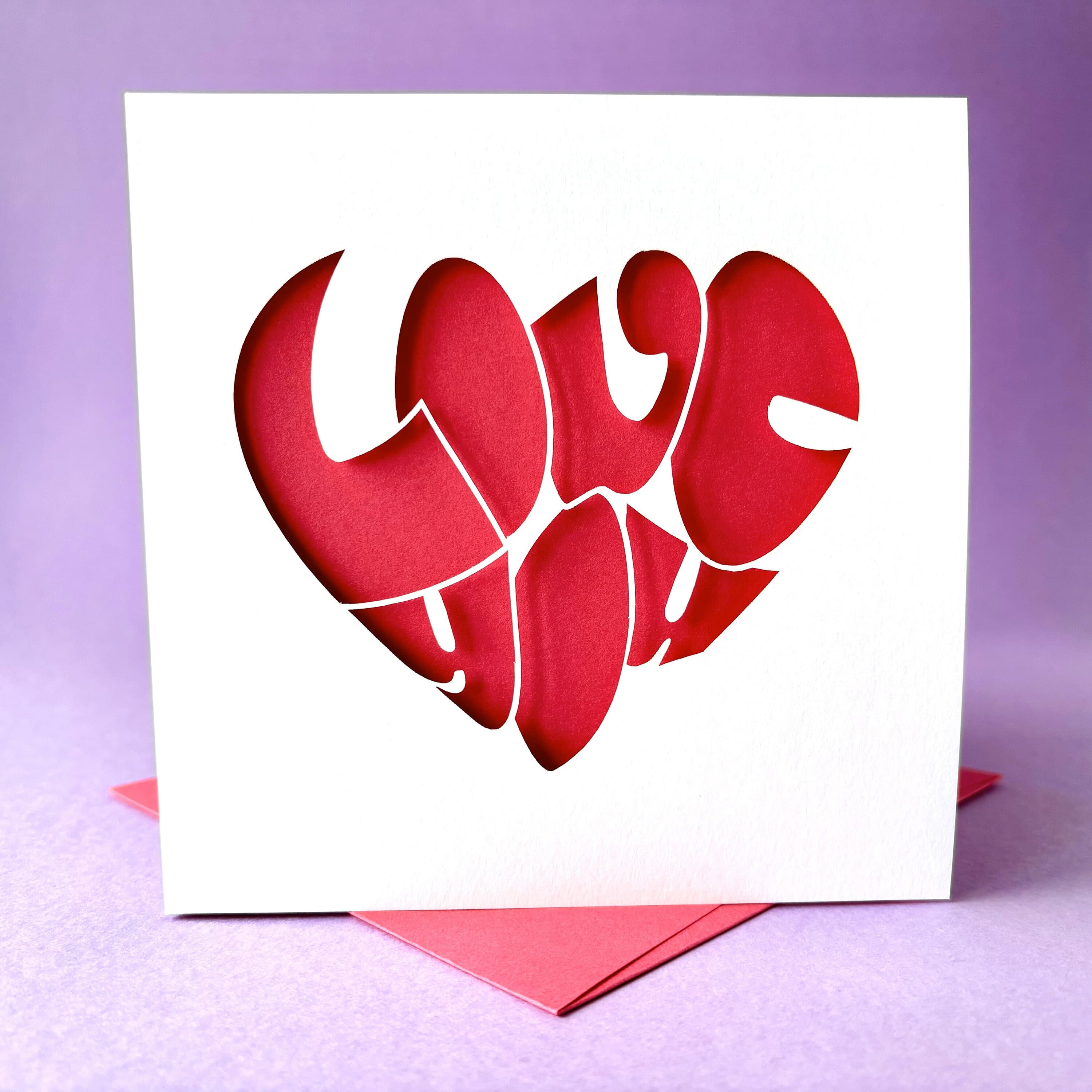 White square laser cut anniversary or Valentine's card. Featuring the words 'Love You' in a bold bubble font shaped into a heart shape. The card has a hot pink paper insert and is standing on a colour-matched envelope. The card is standing against a pale purple background.