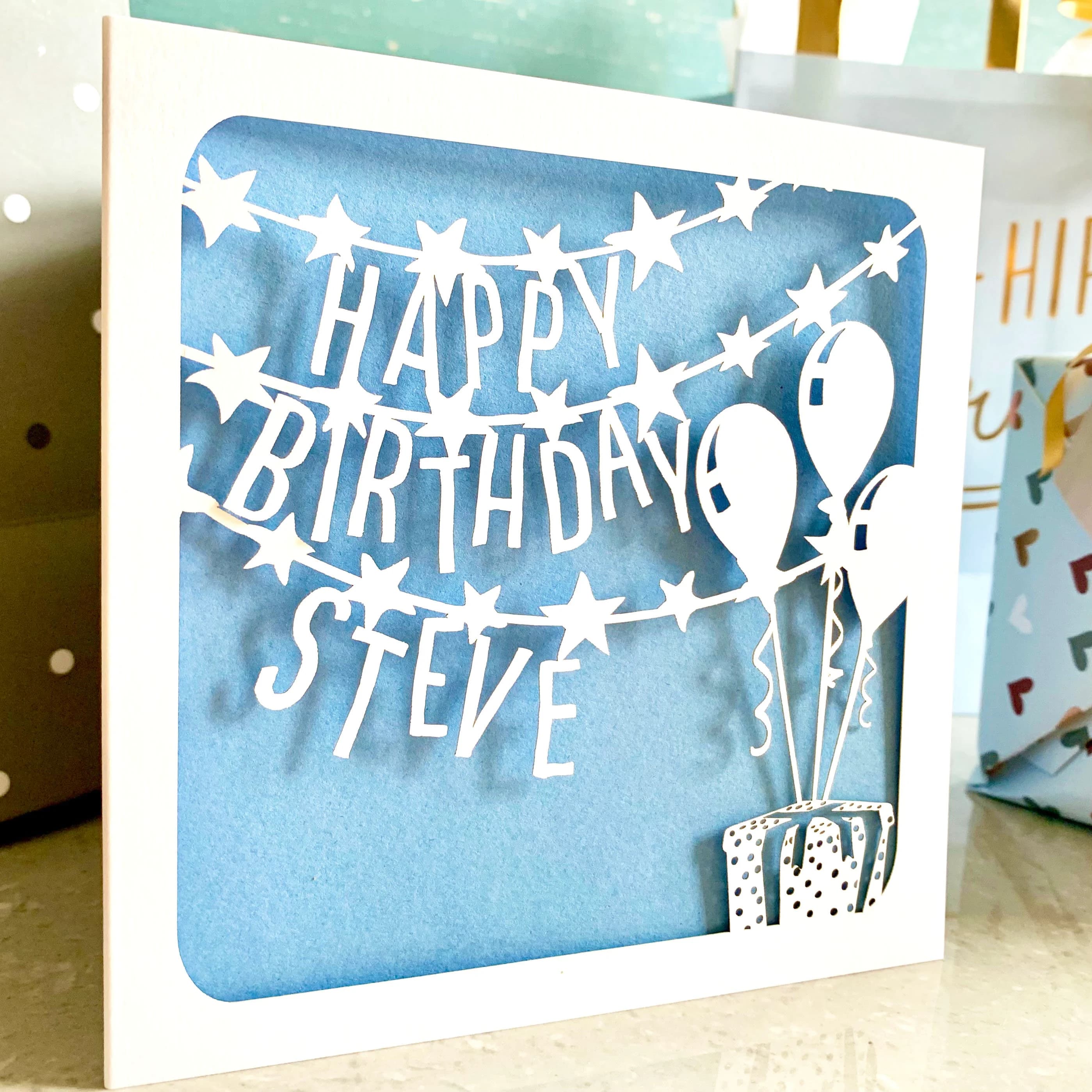 White square laser cut birthday card. Featuring star-decorated banners running across the page, with the words 'Happy', 'Birthday' and a personalised name on each line. The design is finished with a present in the bottom right corner, with three balloons floating above it. The card has a pale blue paper insert and is standing in a celebratory setting, with gifts and gift bags visible behind.