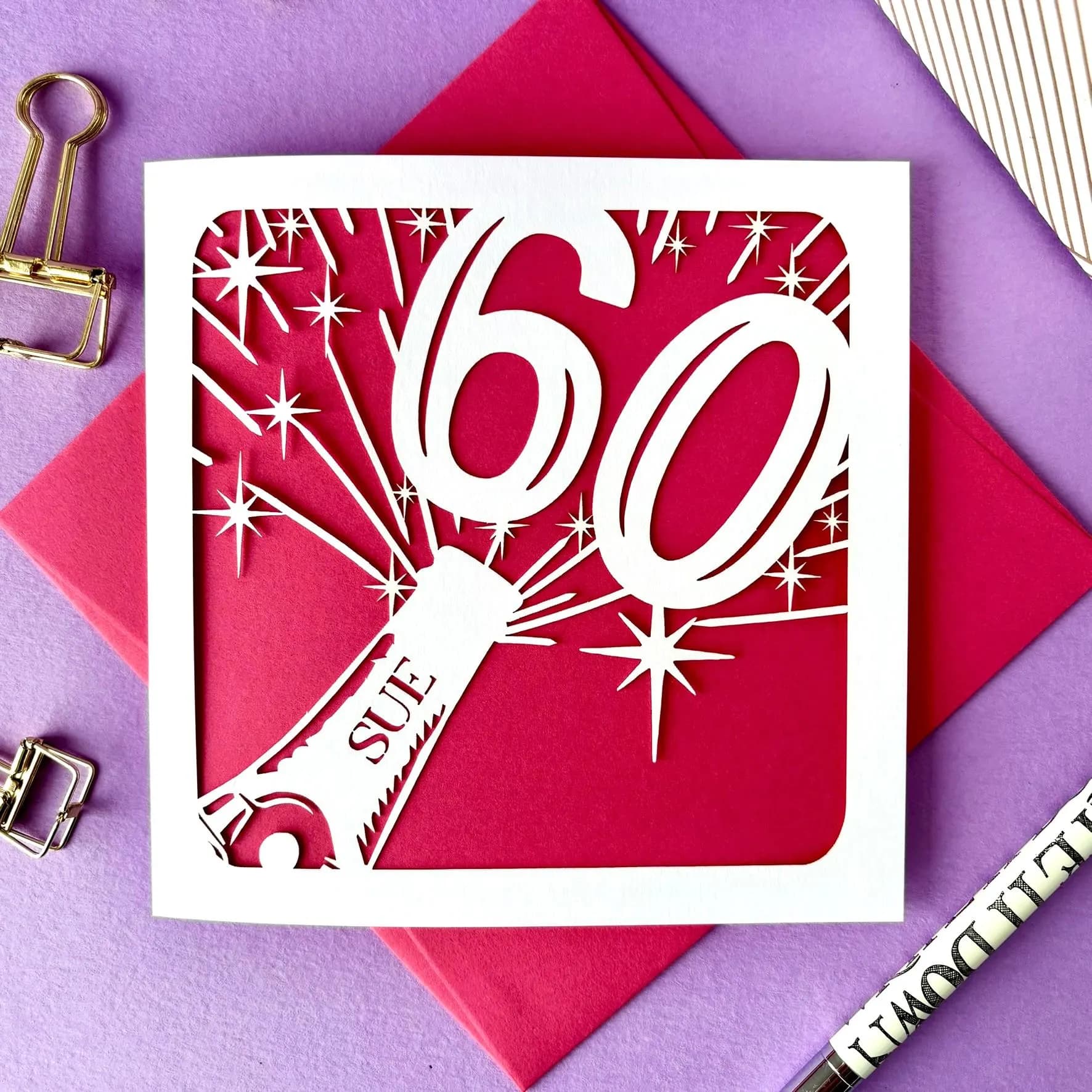 White square laser cut 60th birthday card. Featuring the silhouette of a champagne bottle exploding with a bold number 60 coming out of the top. The card is personalised with a name on the neck of the bottle. The card has a hot pink paper insert and is lying on a colour-matched envelope. The card is lying against a pale purple background with stationery visible surrounding.