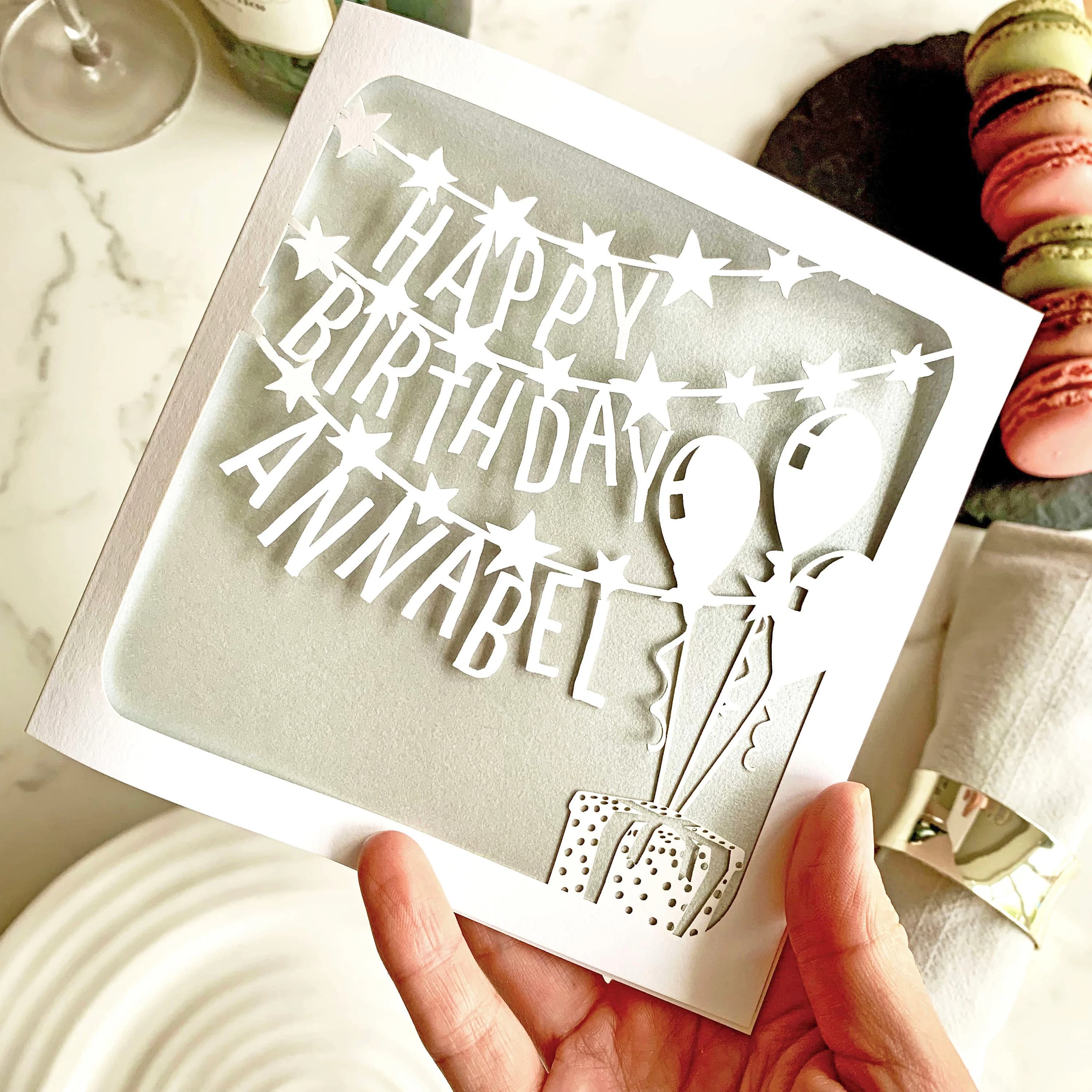 White square laser cut birthday card. Featuring star-decorated banners running across the page, with the words 'Happy', 'Birthday' and a personalised name on each line. The design is finished with a present in the bottom right corner, with three balloons floating above it. The card has a silver paper insert and is being held above a table set for a celebration, with wine glasses and cakes visible behind.