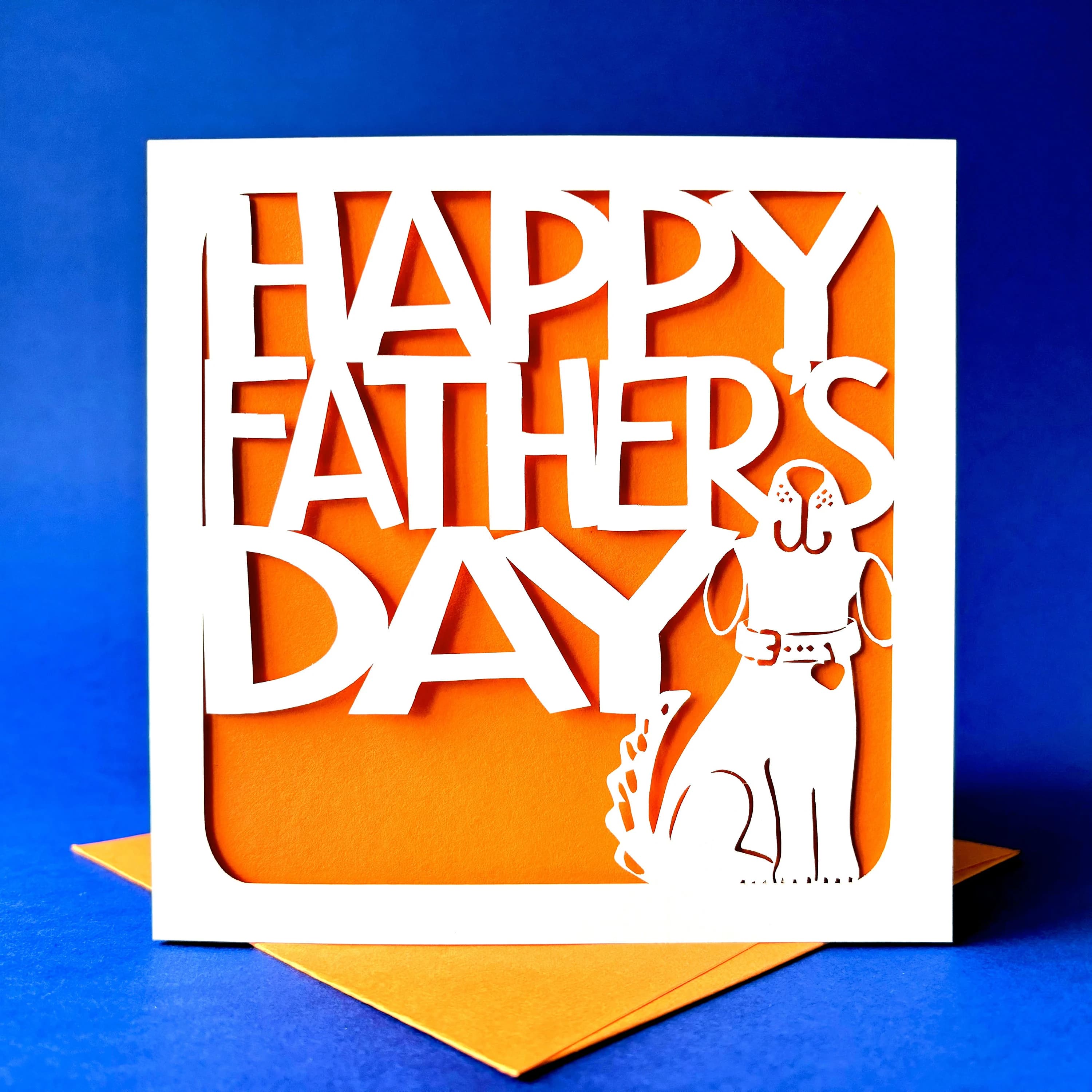 White square laser cut Father's Day card. Featuring a cute dog outline, looking up and wagging his tail. Behind the words 'Happy Father's Day' are cut in a bold font. The card has an orange paper insert and is standing on a colour-matched envelope. The card is standing against a dark blue background.