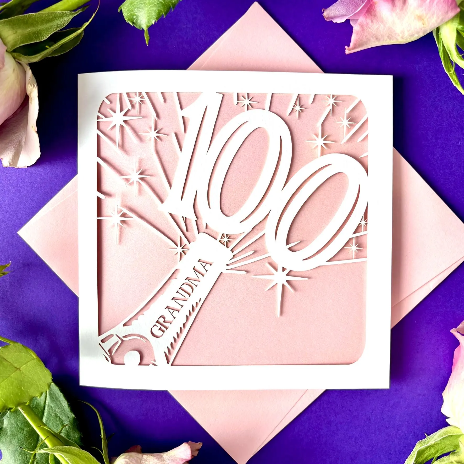 White square laser cut 100th birthday card. Featuring the silhouette of a champagne bottle exploding with a bold number 100 coming out of the top. The card is personalised with a name on the neck of the bottle. The card has a pale pink paper insert and is lying on a colour-matched envelope. The card is lying against a purple background with flowers visible surrounding.