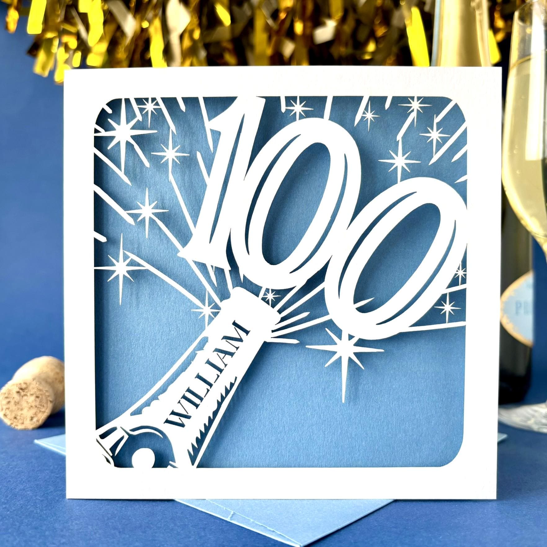 White square laser cut 100th birthday card. Featuring the silhouette of a champagne bottle exploding with a bold number 100 coming out of the top. The card is personalised with a name on the neck of the bottle. The card has a pale blue paper insert and is standing on a colour-matched envelope. The card is standing against a dark blue background with champagne and gold streamers visible behind.