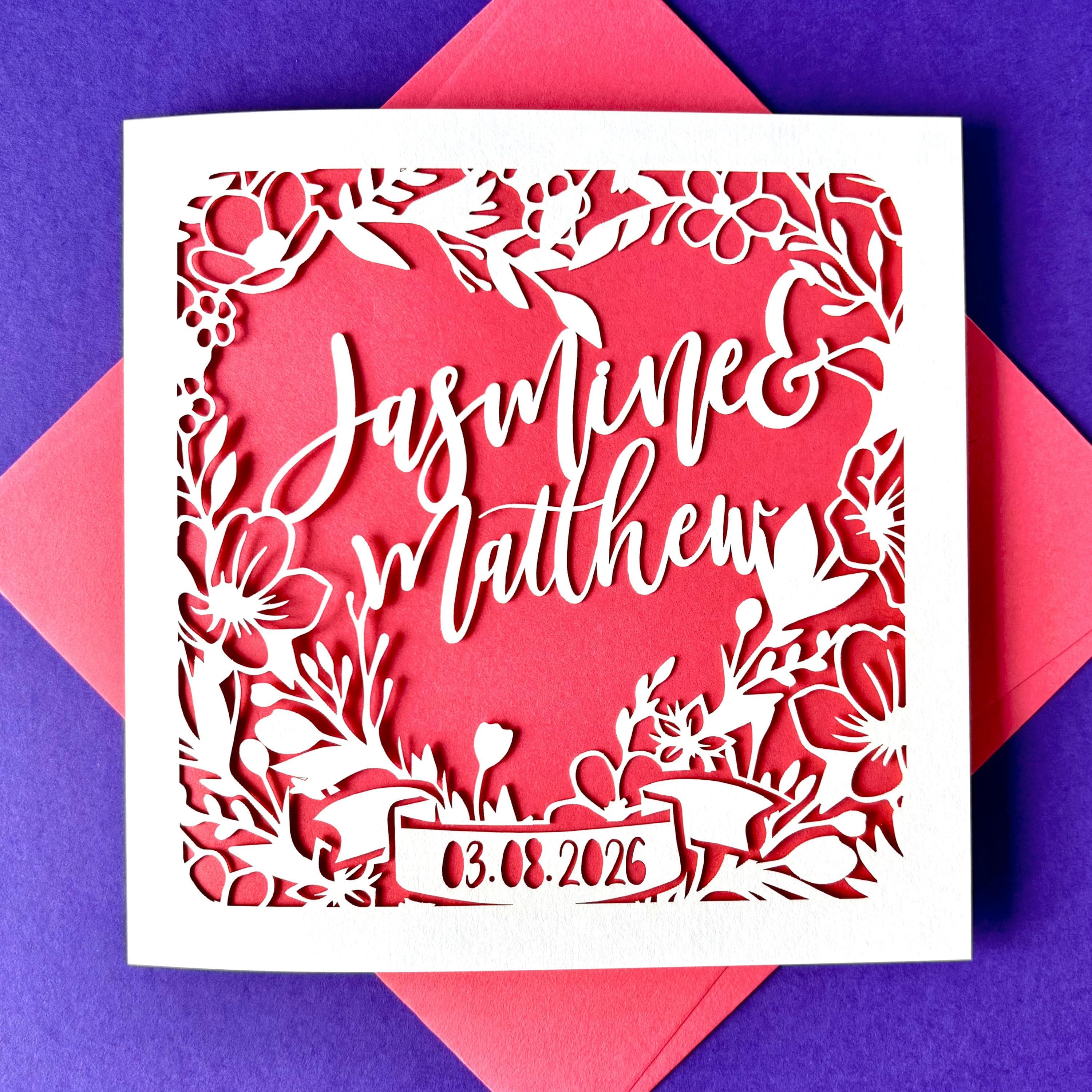 Square white laser cut wedding card featuring a heart shaped floral wreath. The centre of the wreath is personalised with two names in an elegant script font. The base of the wreath features a ribbon banner, with a date showing across it. The card has a hot pink paper insert, and is lying on a colour-matched envelope. The card is lying against a purple background.