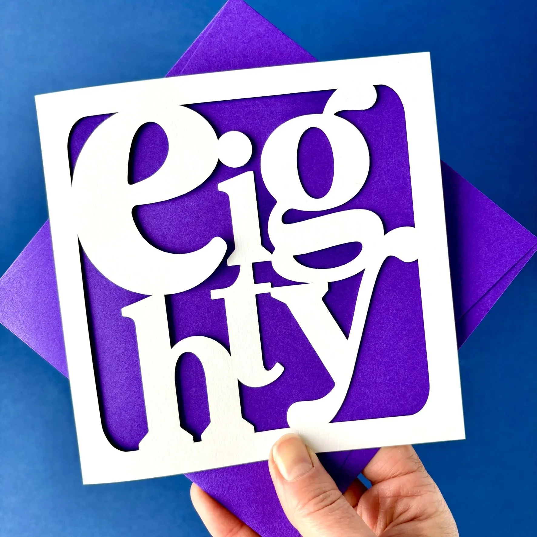 White square laser cut 80th birthday card. Featuring the number 80 written in bold modern text in a fun layout. The card has a purple paper insert and is being held on a colour-matched envelope. The card is being held against a dark blue background.