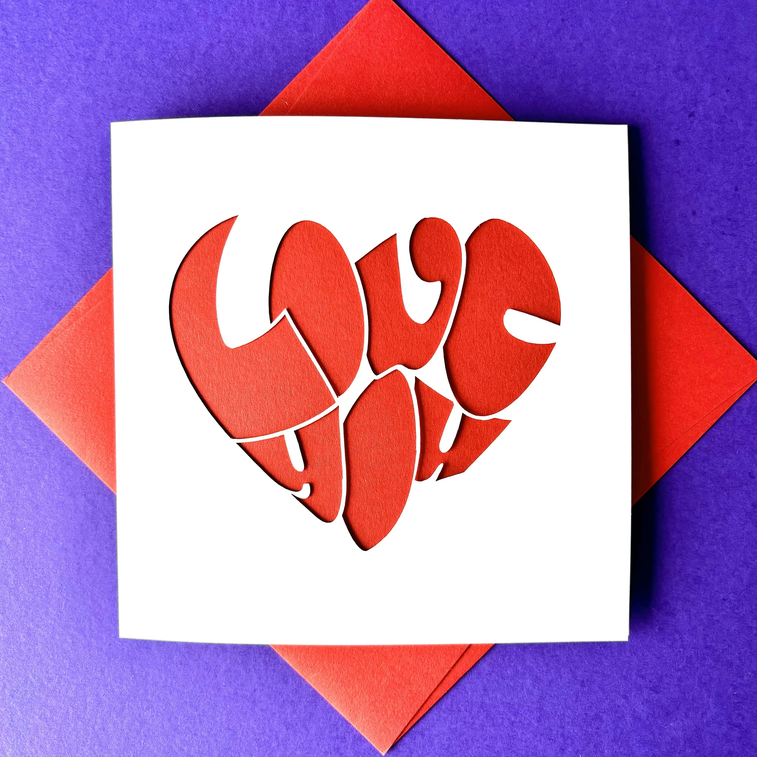 White square laser cut anniversary or Valentine's card. Featuring the words 'Love You' in a bold bubble font shaped into a heart shape. The card has a red paper insert and is lying on a colour-matched envelope. The card is lying against a purple background.