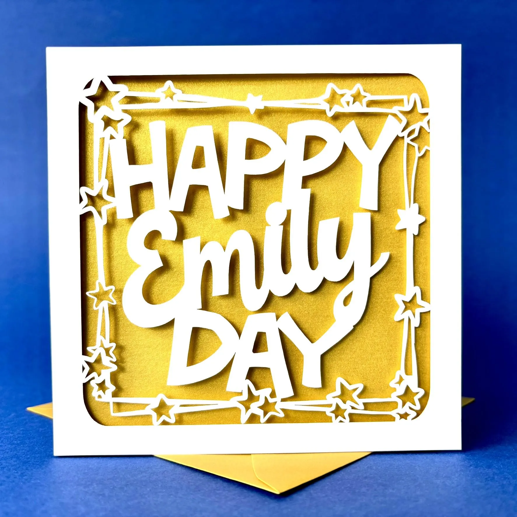 White square laser cut personalised birthday card. Featuring the words 'Happy [NAME] Day', in a bold, playful cut‑out font. The words are framed by a stylish border scattered with star outlines. The card has a gold paper insert and is standing on a colour-matched envelope. The background is dark blue.