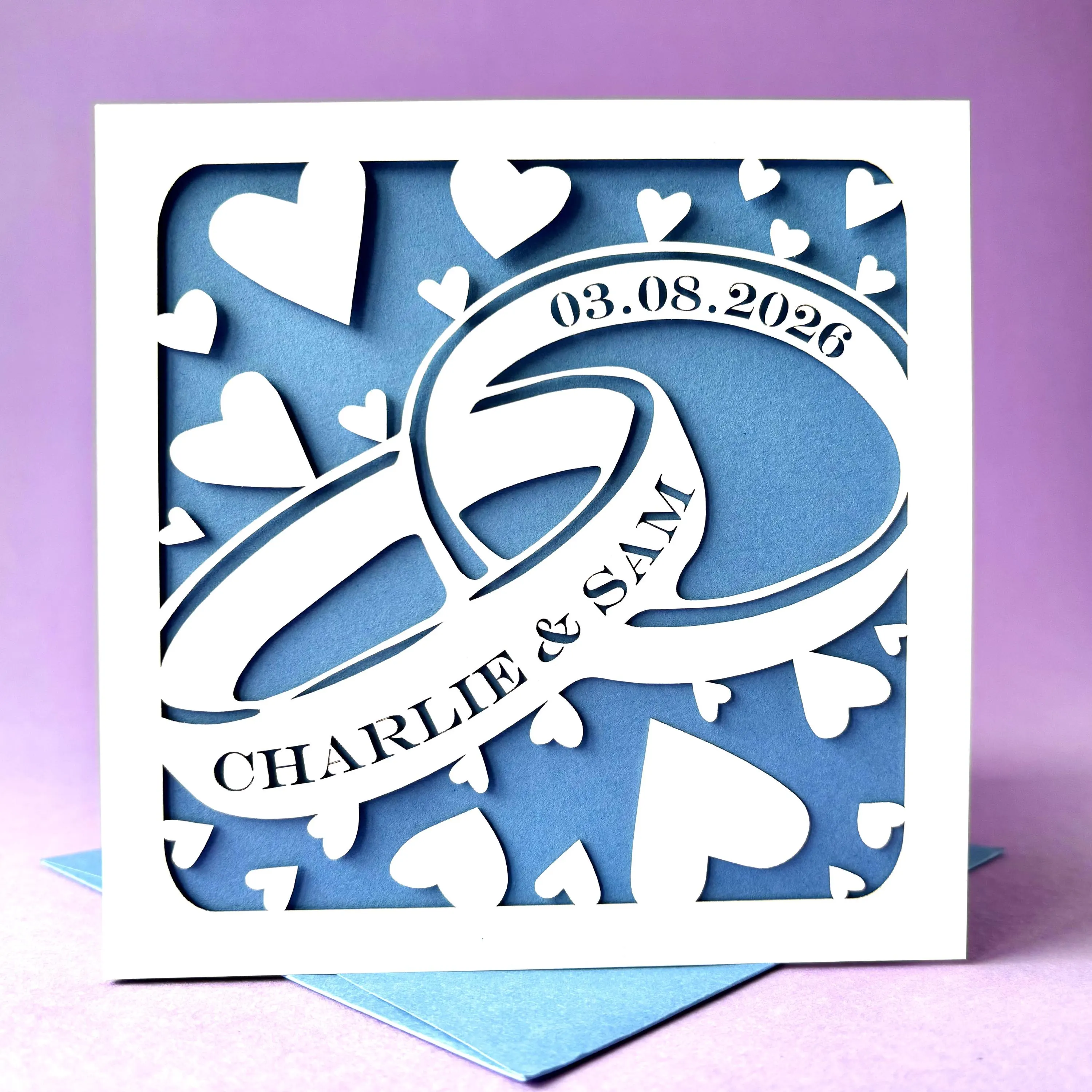 Square white laser cut greetings card featuring two entwined rings surrounded by hearts. The front ring has been personalised with two names, and the second with a date, in a traditional serif font. The wedding card has a pale blue paper insert and is standing on a matching envelope. It is stood against a light purple background.