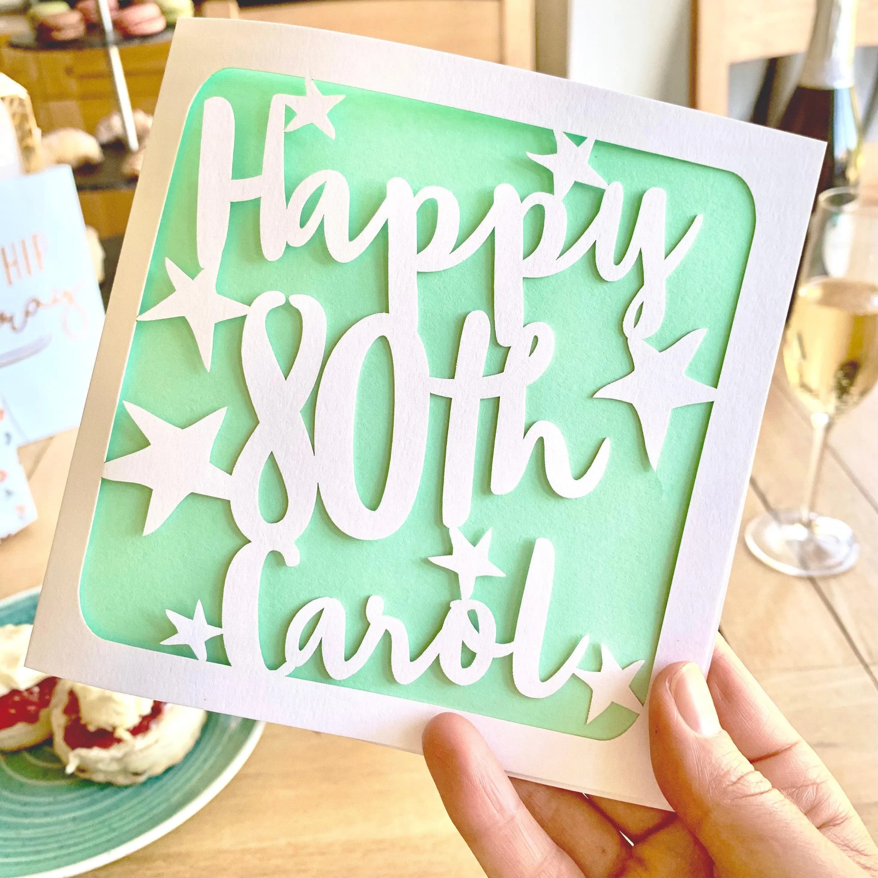 White square laser cut 80th birthday card. Featuring the words ‘Happy 80th’ cut in a bold script font across the card, which has been personalised with a name in the last line. Hand-drawn stars surround the text. The card has a mint green paper insert and is being held up in front of a celebratory afternoon tea scene.