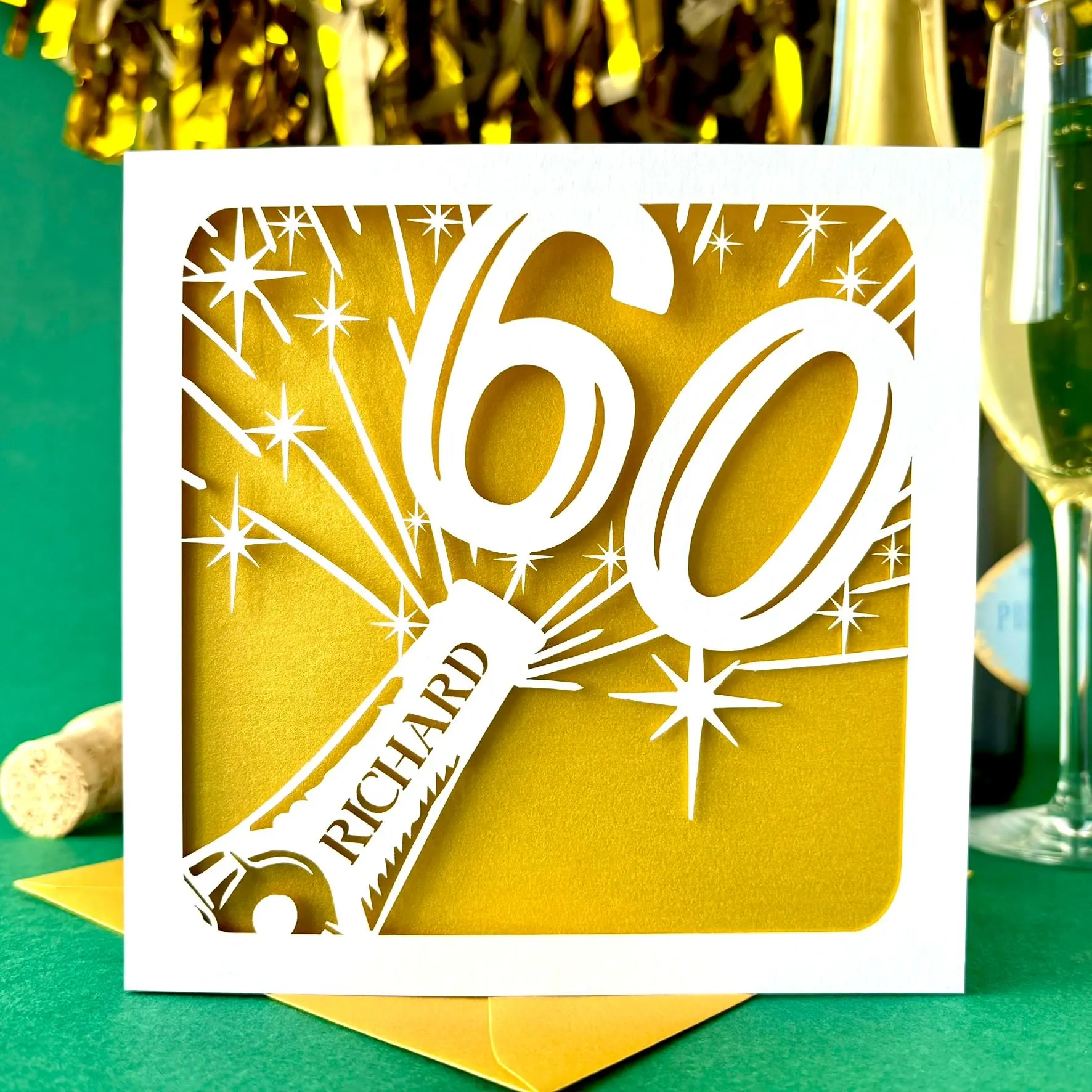 White square laser cut 60th birthday card. Featuring the silhouette of a champagne bottle exploding with a bold number 60 coming out of the top. The card is personalised with a name on the neck of the bottle. The card has a gold paper insert and is standing on a colour-matched envelope. The card is standing against a dark green background with champagne and gold streamers visible behind.