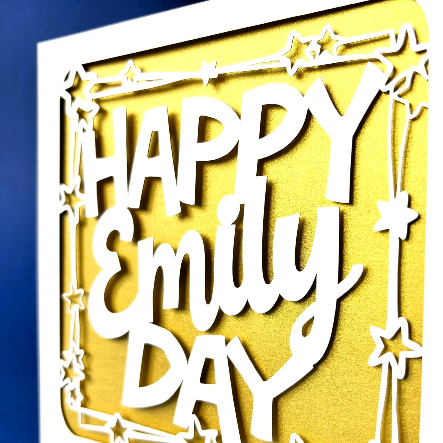 Close up of a white square laser cut personalised birthday card. Featuring the words 'Happy [NAME] Day', in a bold, playful cut‑out font. The words are framed by a stylish border scattered with star outlines. The card has a gold paper insert and the background is dark blue.