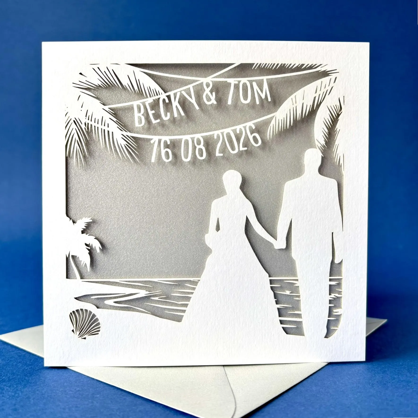 White square laser cut wedding card featuring the silhouette of a bride and groom wedding couple on a beach, surrounded by palm trees. Above the couple the card has been personalised with bunting which shows their name and the date of the wedding. The card has a silver paper insert and is standing on a colour matched envelope. It is stood against a dark blue backdrop.