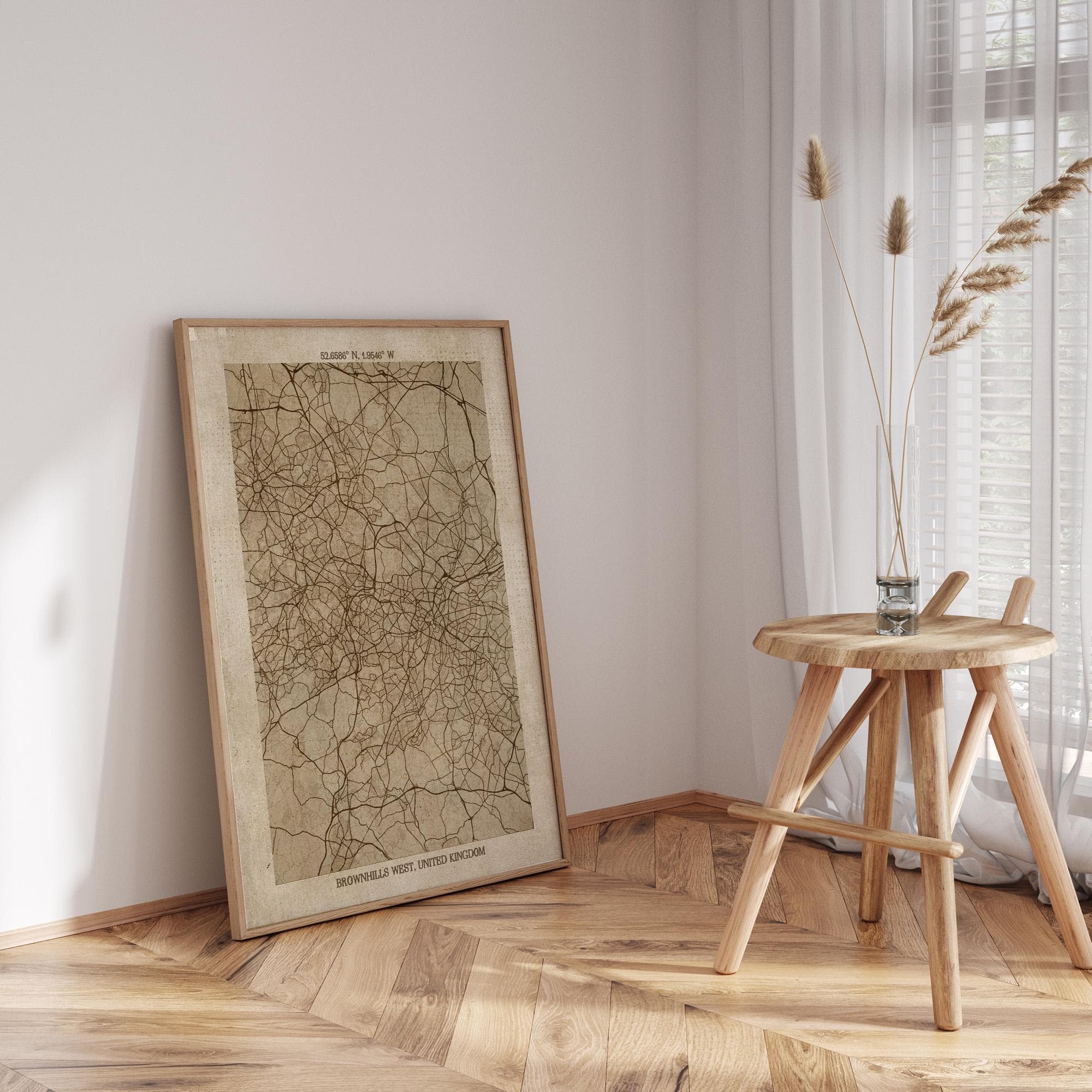 a framed map of the United Kingdom, a wooden stool, and a vase with dried reeds, all placed in a corner of a room with a wooden floor.