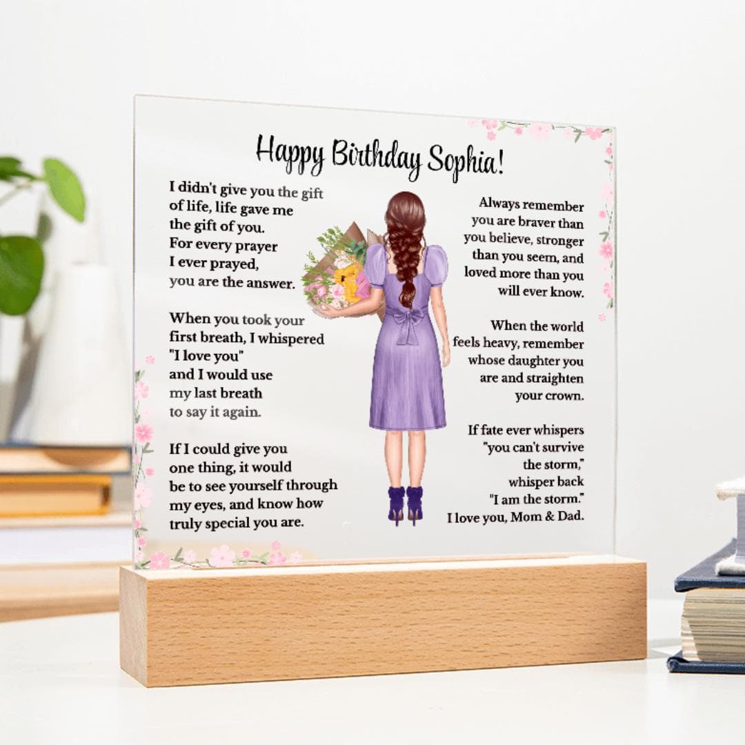 May Include: A personalized acrylic plaque displayed on a wooden LED base in a home setting. The custom design features a birthday message, pink floral accents, and an illustrated girl with braided auburn hair wearing a purple dress and holding a bouquet. A cactus and decorative vase are blurred in the background.