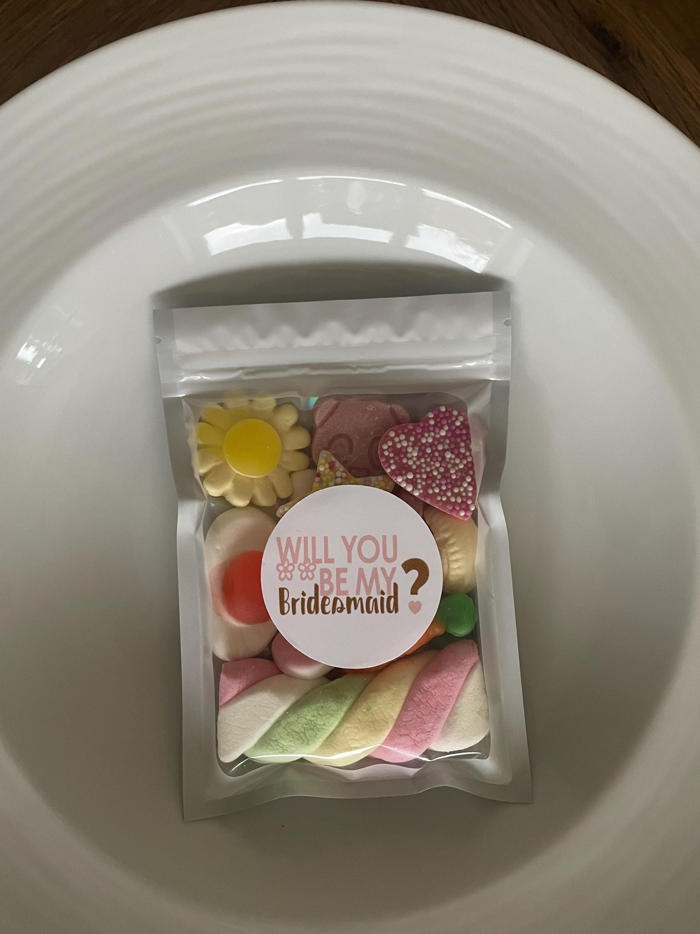 May include: A clear bag of sweets, including a yellow flower, a pink heart, and a rainbow-coloured marshmallow. The bag has a round sticker with the text "Will you be my Bridesmaid?" in pink.
