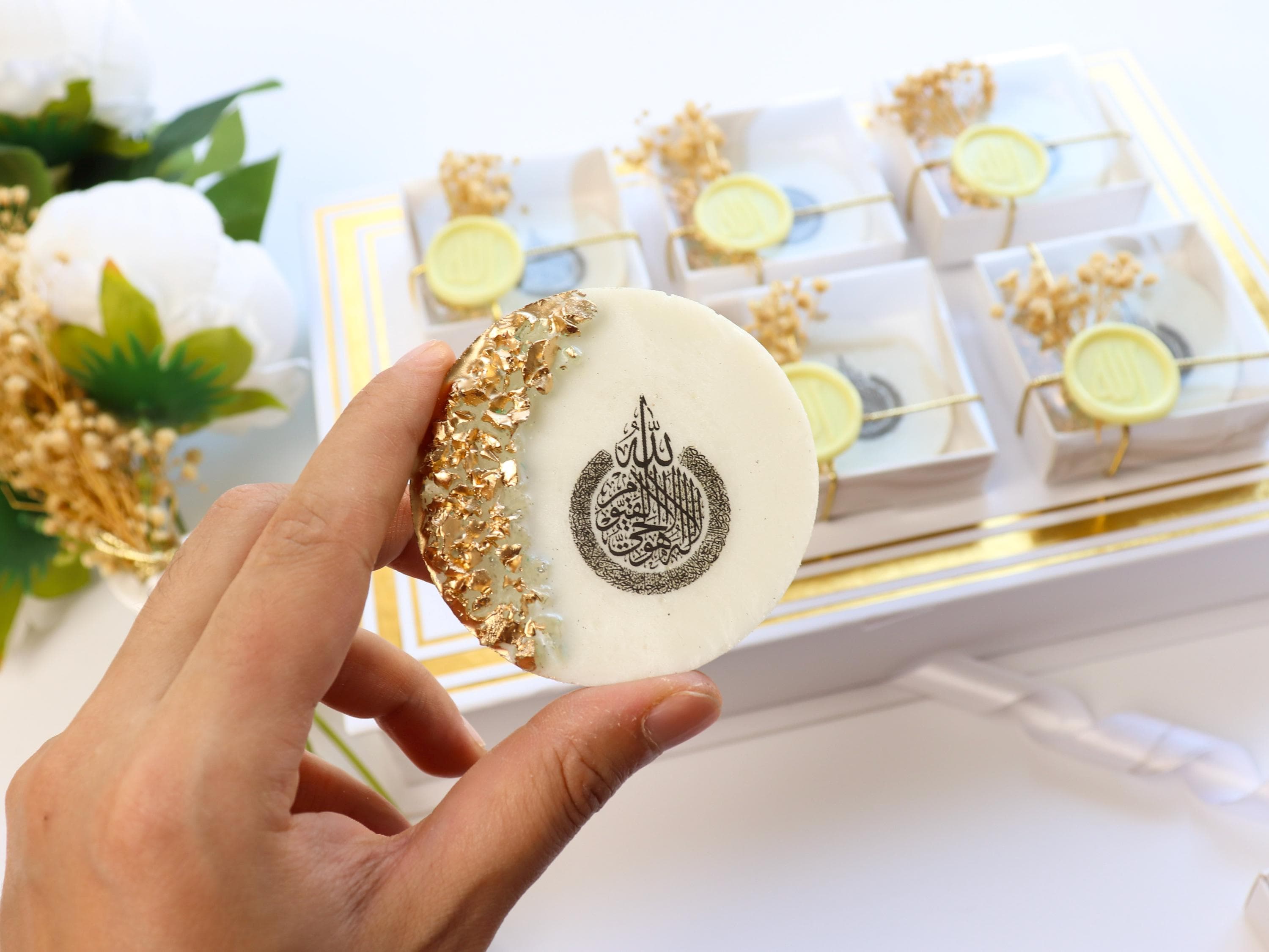 May include: A round, white favor with gold leaf accents and a black Arabic calligraphy design is held in a hand. The favor is part of a set of similar gifts in a white box, decorated with gold and yellow wax seals.