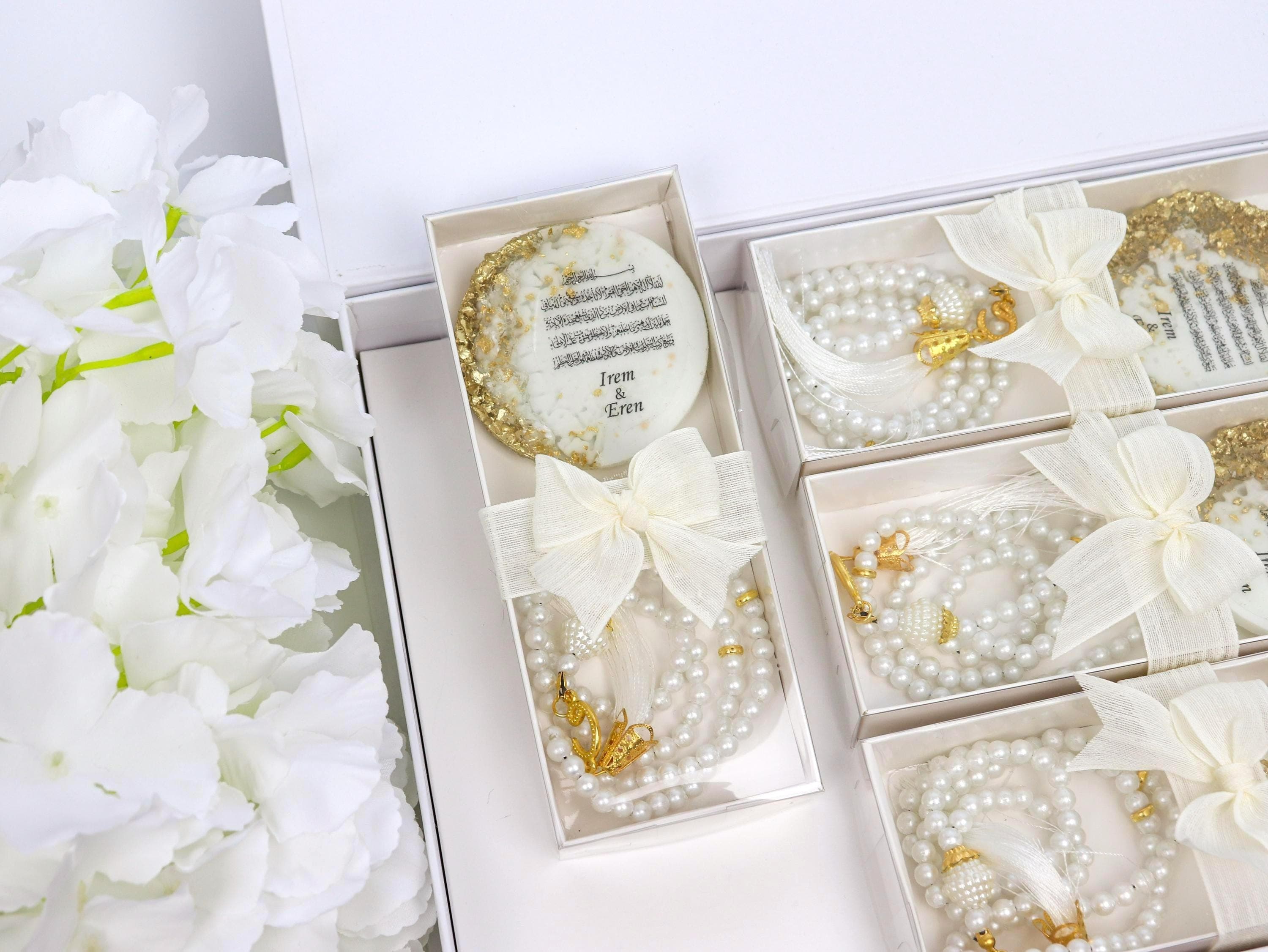 May include: White gift boxes with pearl necklaces, decorative accents, and a round, gold-rimmed item with text. Each box is tied with a cream-colored bow. The overall theme is elegant and celebratory.