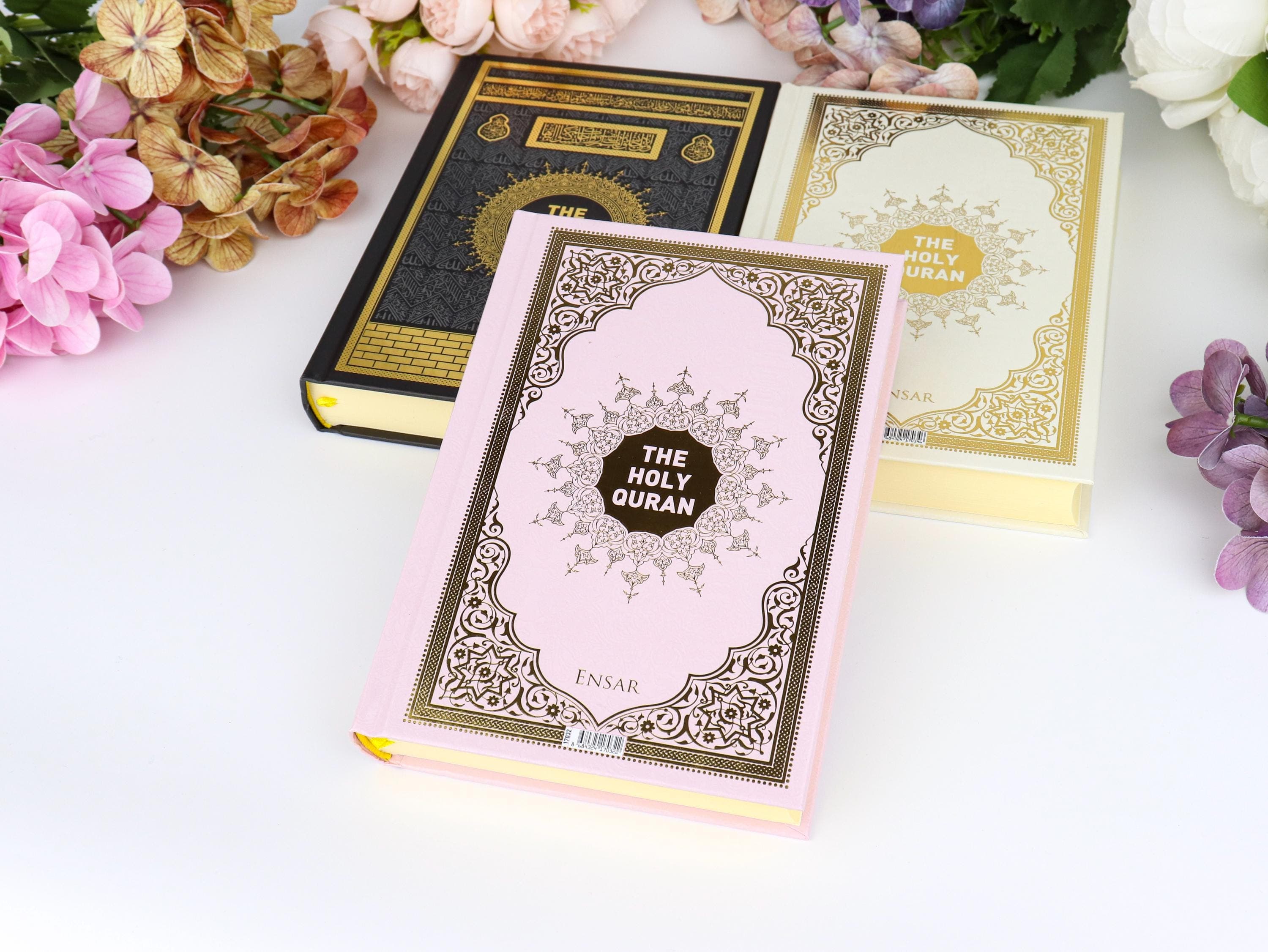 May include: Three copies of "The Holy Quran" are displayed, each with ornate gold and black or pink covers. The central book is pink with gold accents and the title in a black circle. Floral arrangements in pink, brown, and purple surround the books.