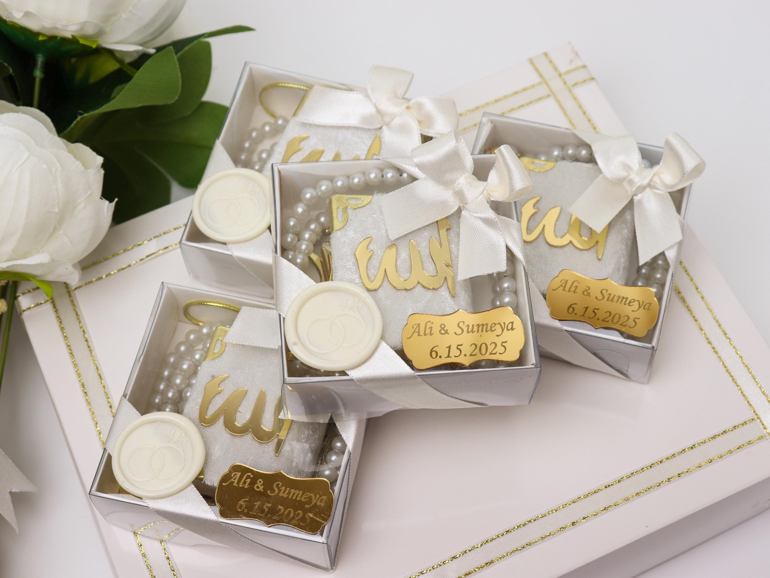 May include: White gift boxes with gold accents, each containing a pearl strand, a decorative item with gold script, and a small gold tag with names and a date. The boxes are tied with white satin bows.