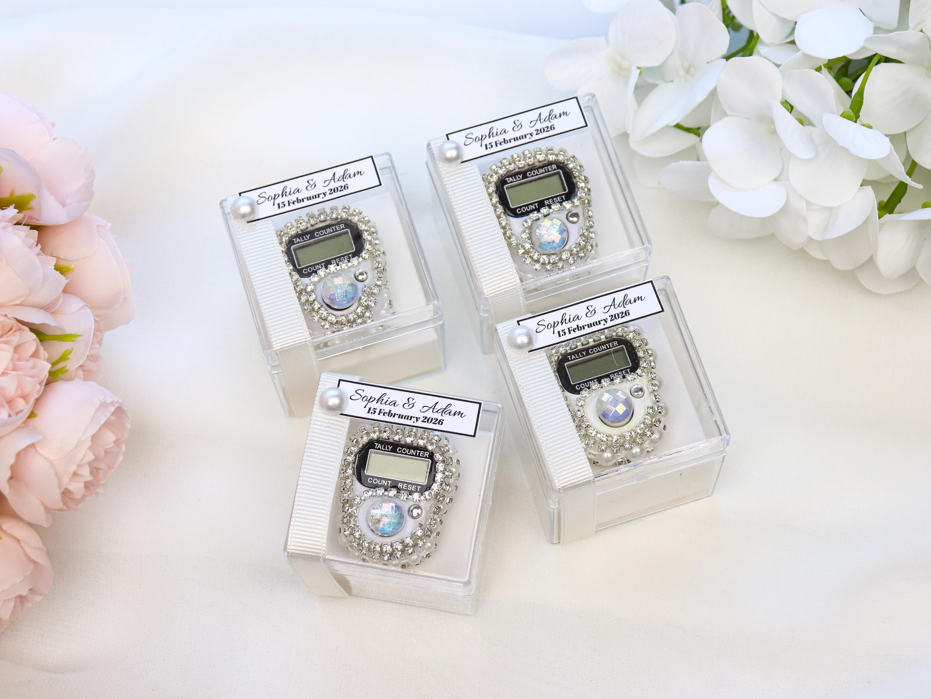 May include: Four silver tally counters in clear boxes, each with a digital display and embellished with rhinestones. Each box has a label that reads "Sophia & Adam 15 February 2026". White ribbon and pearl accents complete the presentation.
