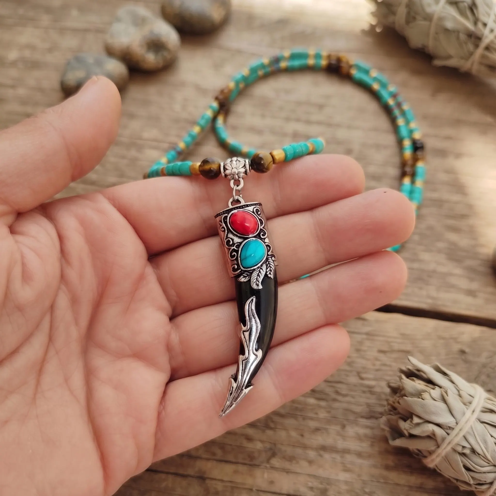 Tribal tusk necklace for men held in hand showing carved antique silver amulet cap with coral red and blue stones on beaded strand