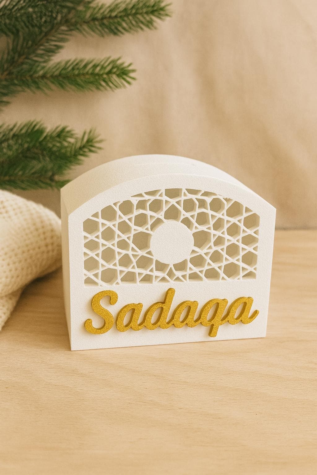 May include: A white decorative box featuring a geometric lattice pattern on the front. The word "Sadaqa" is written in gold script. The box has a curved top and is likely used for storage or display, possibly for gifts.