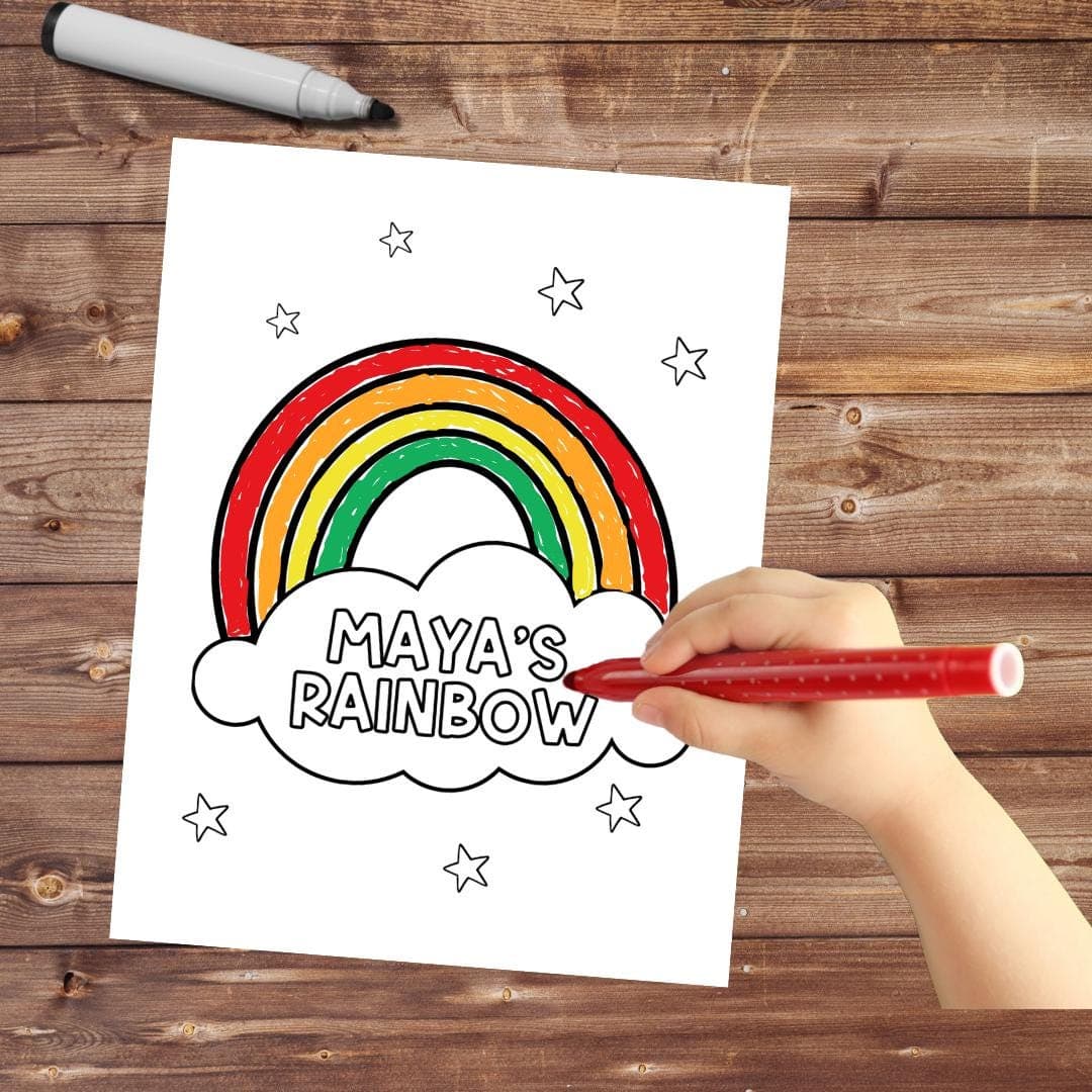 Child coloring a personalized rainbow-themed coloring page titled "Maya's Rainbow" with a red marker on a wooden table, featuring a colorful rainbow and stars.