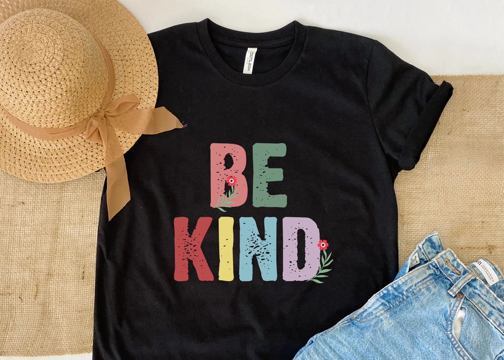 May include: A black t-shirt with the words 'BE KIND' printed in colorful letters with a distressed look. The letters are outlined in black and have small floral accents. The shirt is laid flat on a textured surface.