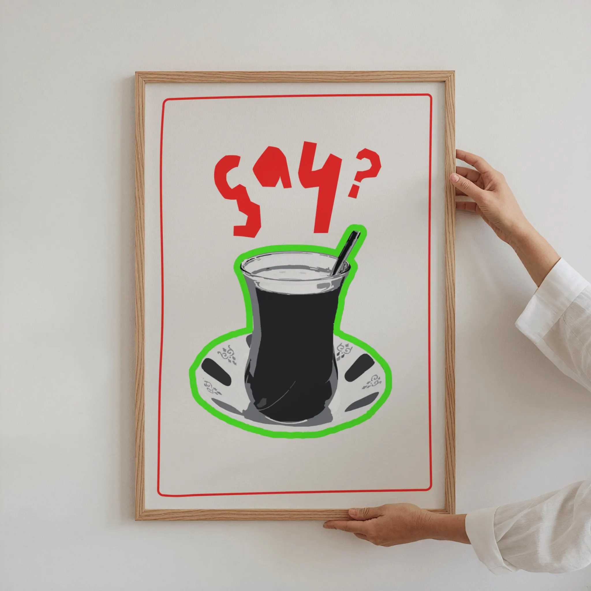 A framed art print featuring a stylized illustration of a Turkish tea glass on a saucer, outlined in green. The text "çay?" is in red above the glass. The frame is wood-toned.
