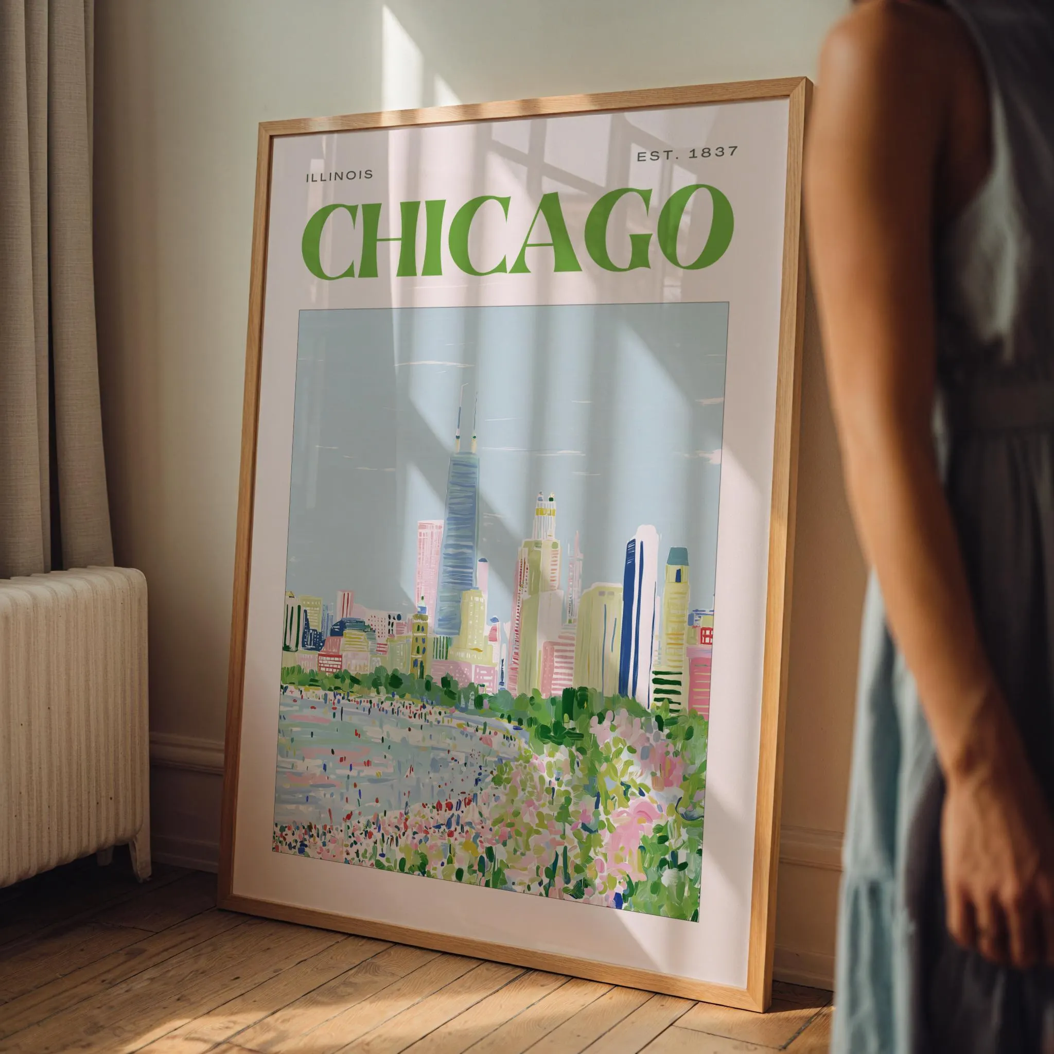 A framed wall art print featuring a stylized illustration of the Chicago skyline. The text reads ILLINOIS, CHICAGO, and EST. 1837 in green. The artwork depicts a city landscape with a light blue sky, colorful buildings, and a park.