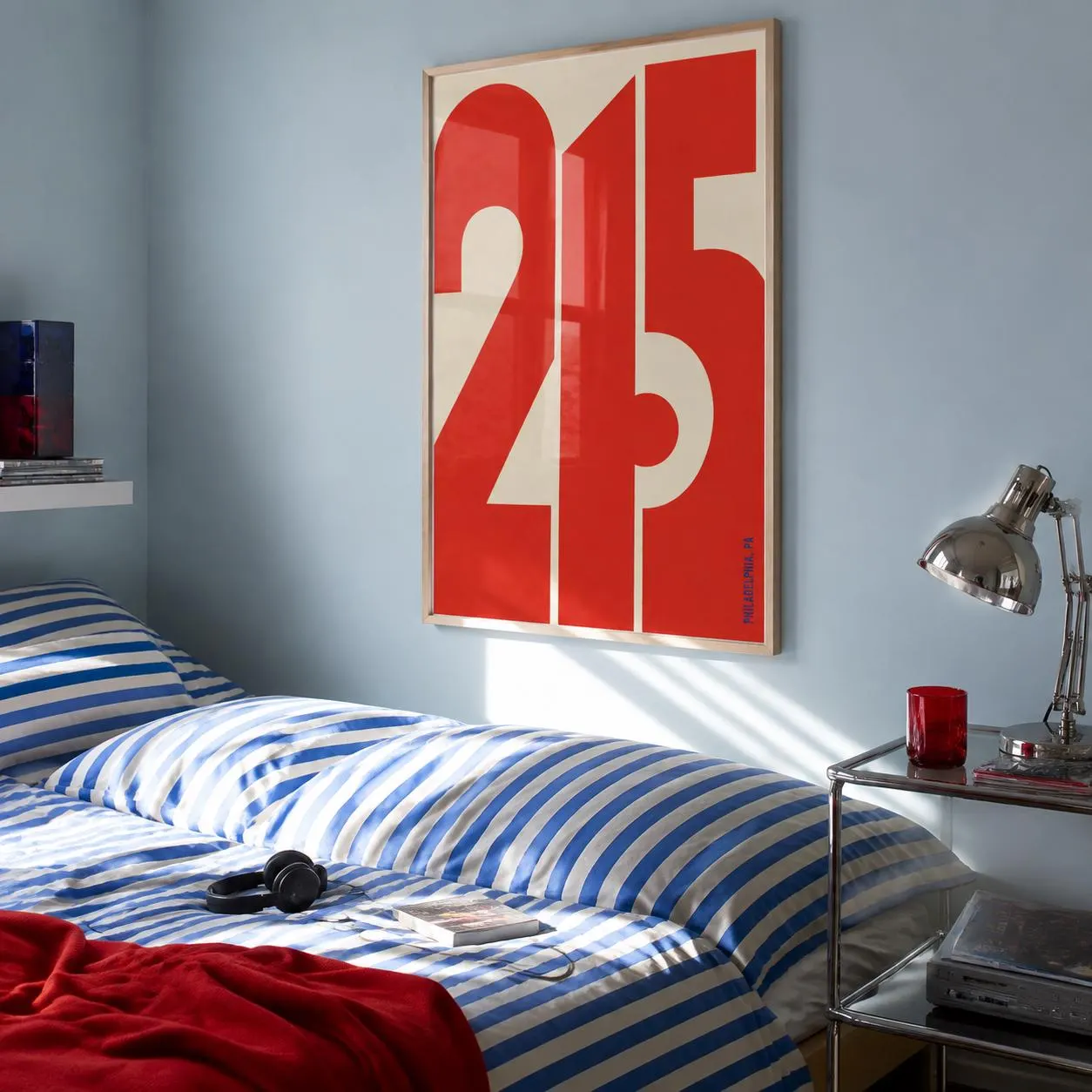 A rectangular wall art print in a light wood frame featuring the large red numbers 215 against a cream background. The print is displayed on a light blue wall above a bed with blue and white striped bedding and a red blanket.