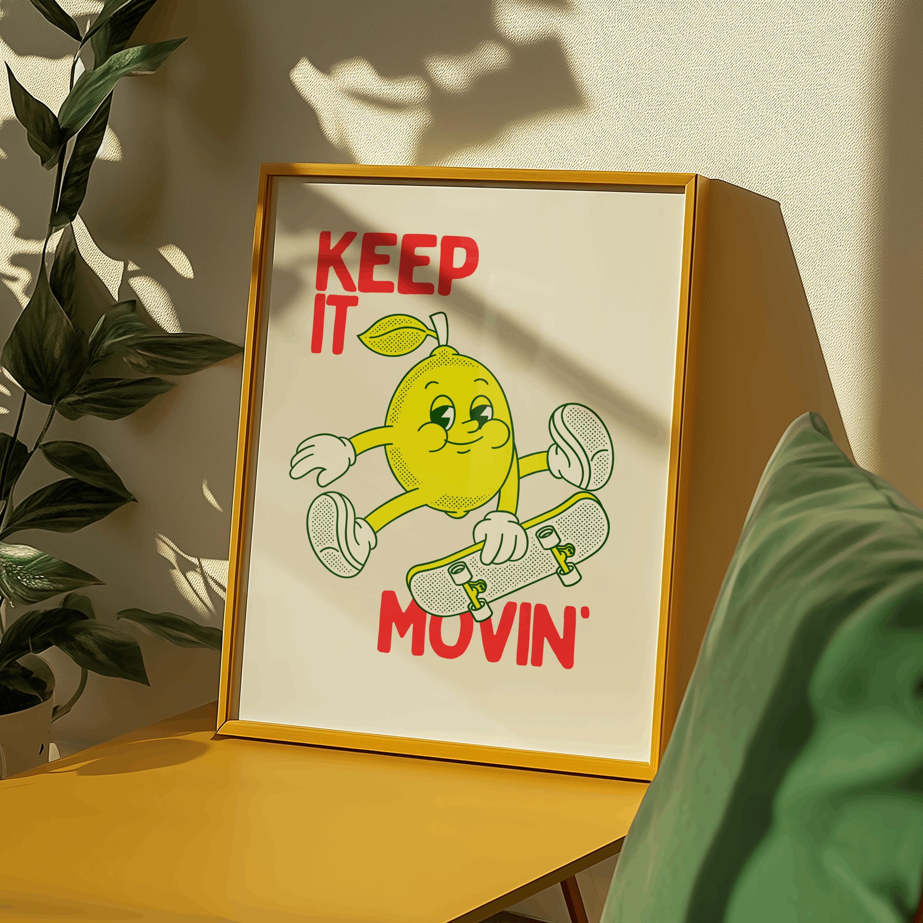 A framed print with a yellow border features a cartoon lemon riding a skateboard. The lemon is yellow with a green leaf and is smiling. The words "KEEP IT MOVIN'" are in red text.