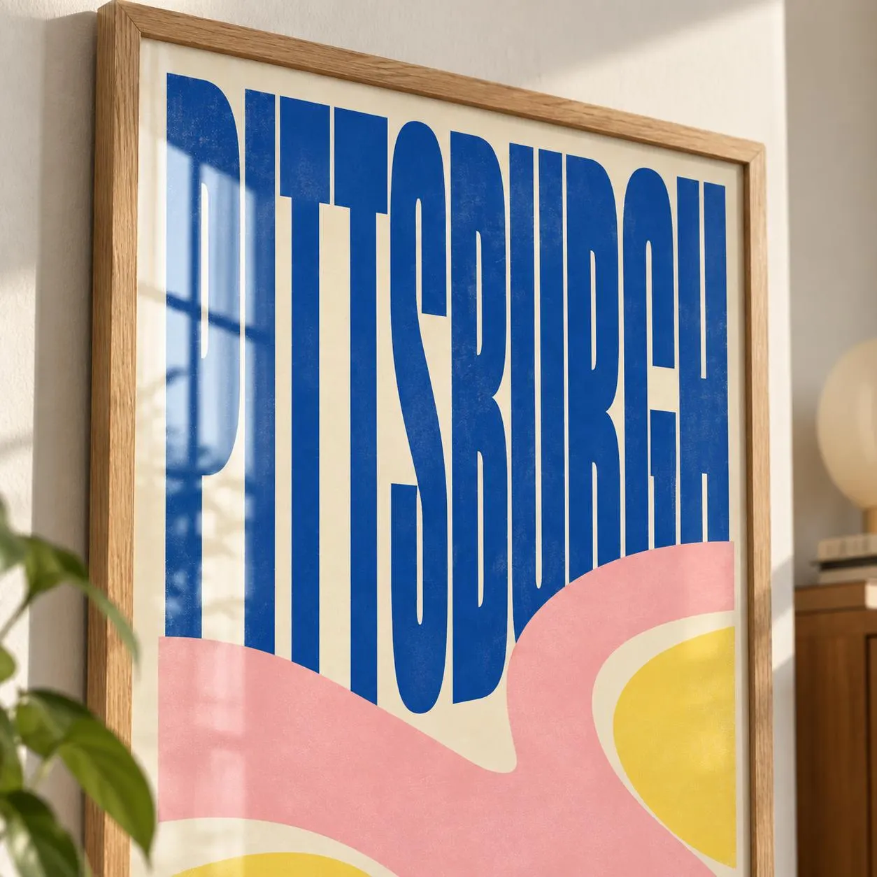 A framed wall art print featuring the word PITTSBURGH in large, bold, blue sans-serif capital letters. The background consists of abstract, wavy, horizontal bands of soft pink and pale yellow colors on a cream-colored base.
