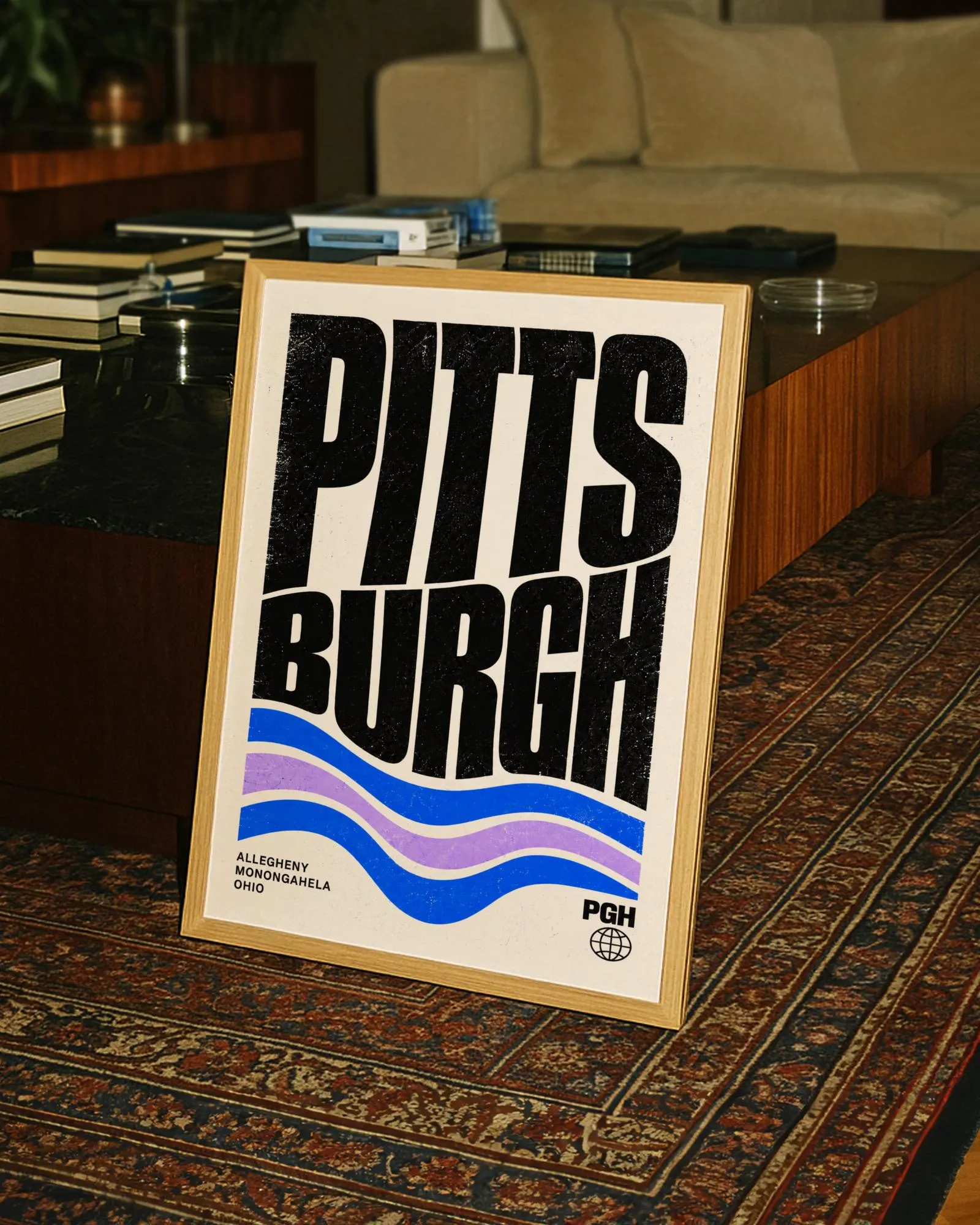 A framed poster featuring the word PITTSBURGH in large black letters. Below the text are three wavy lines in blue and purple. The text ALLEGHENY MONONGAHELA OHIO appears in the bottom left, with PGH and a globe icon in the bottom right.