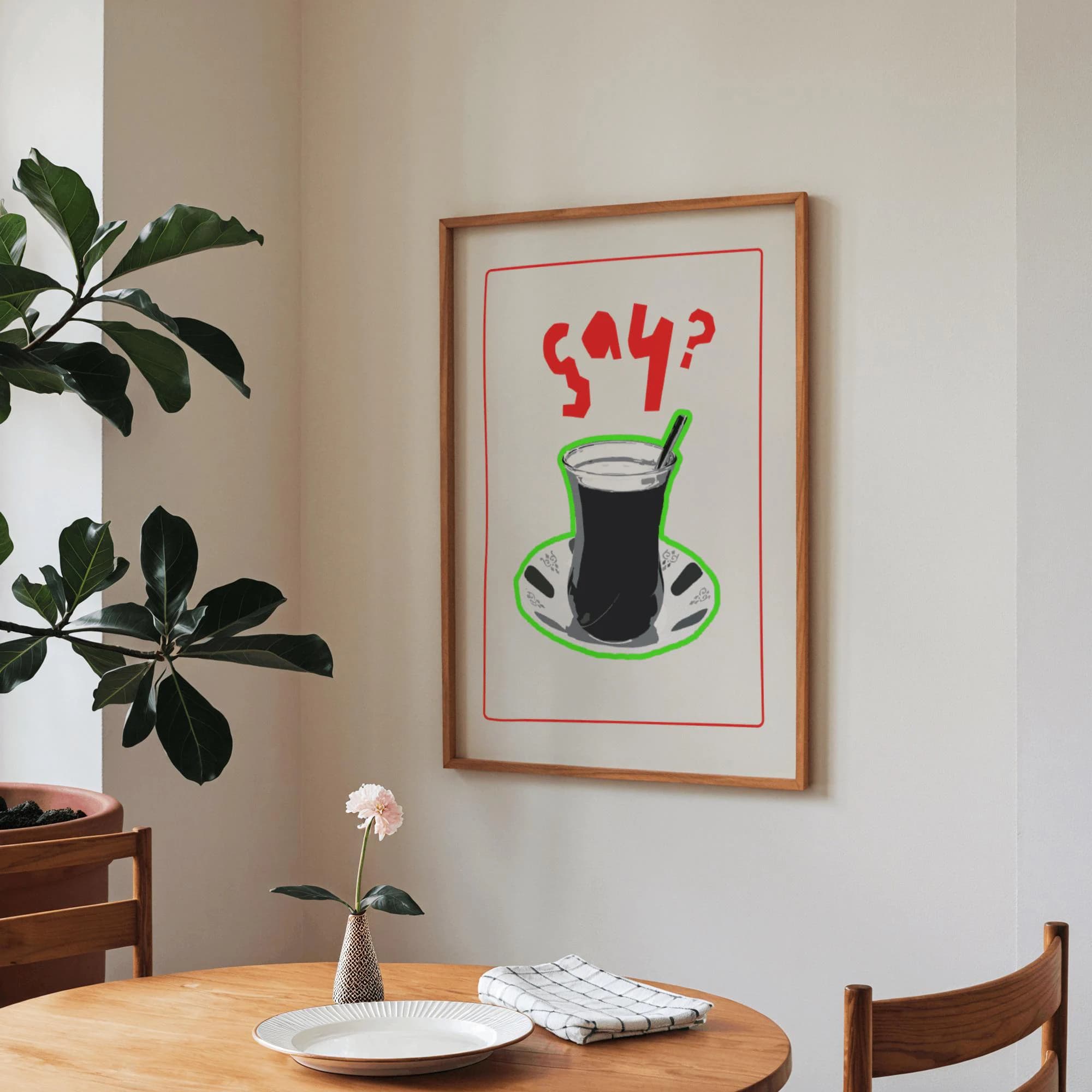 A framed art print featuring a stylized illustration of a Turkish tea glass with a spoon, set on a saucer. The print has a red border and the word "çay?" in red. The artwork is in a wooden frame.