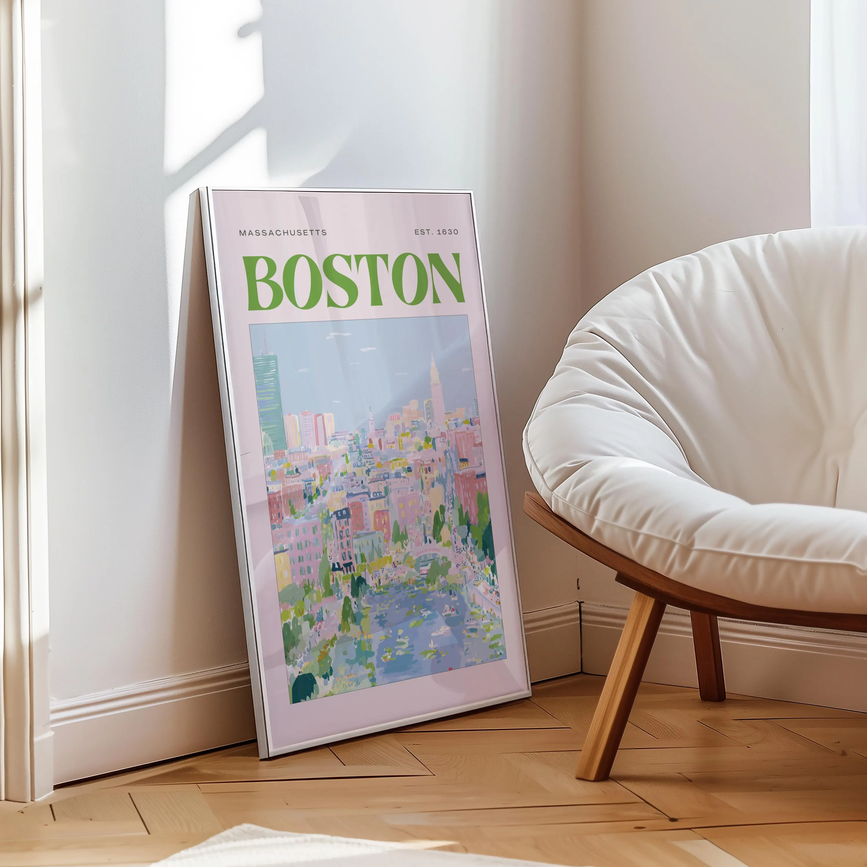A framed art print featuring a colorful illustration of Boston, Massachusetts. The print has a pink background with the city name in large green letters. The artwork is leaning against a wall next to a white chair.