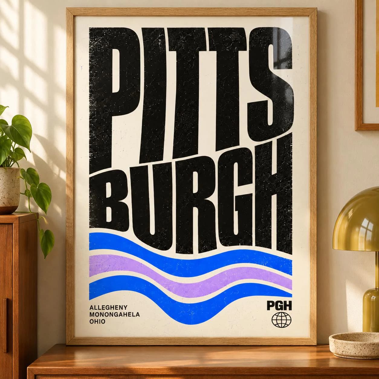 A framed wall art print featuring the word PITTSBURGH in large, distressed black capital letters. Below the text are three wavy horizontal lines in blue and light purple. The text ALLEGHENY MONONGAHELA OHIO and PGH with a globe icon appear.