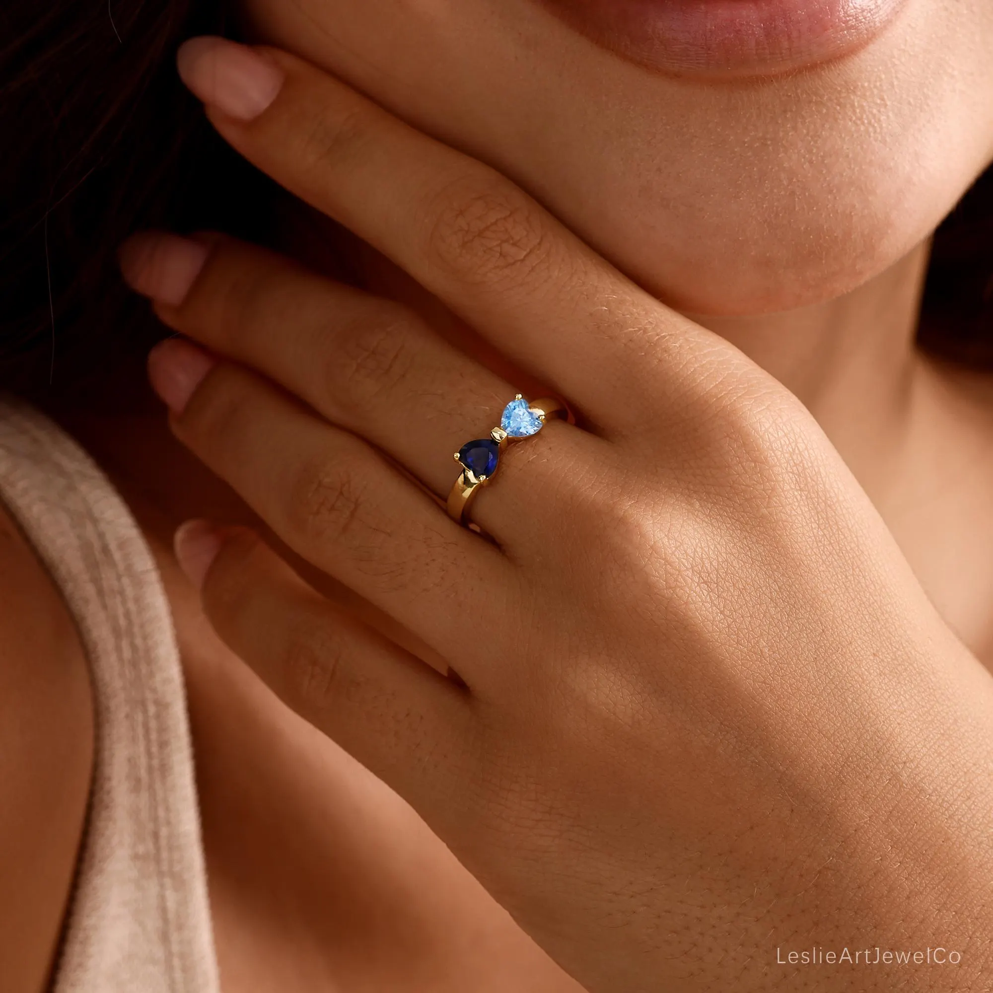 May include: A gold ring featuring two heart-shaped gemstones, one in a deep navy blue and the other in a light sky blue, set side-by-side on a polished gold band. The ring is worn on a finger, showcasing the contrasting blue heart stones.