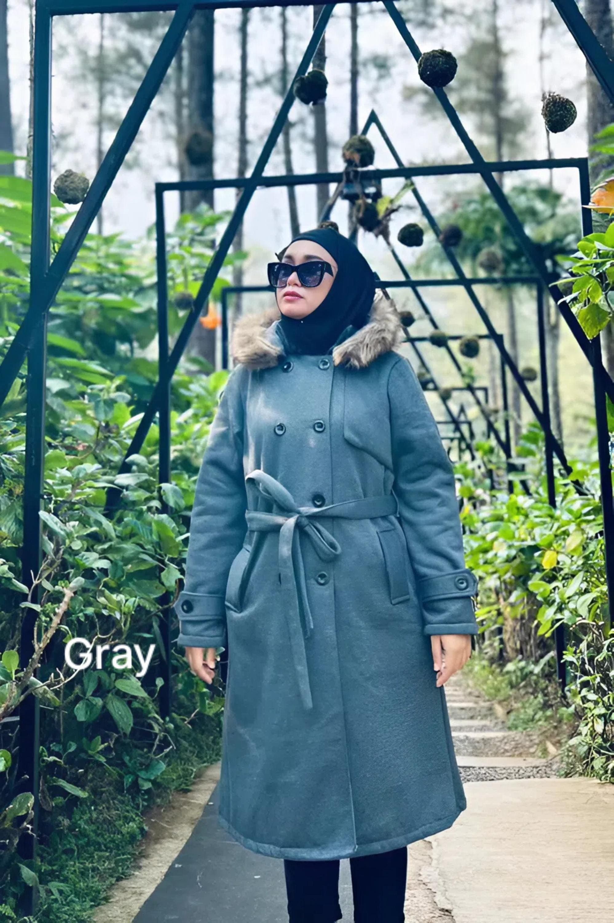 Woman wearing a mocca long fleece winter coat with detachable faux fur hood and belt, standing outdoors in greenery