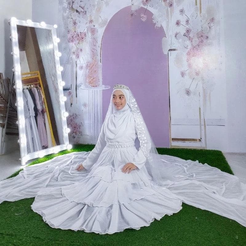 A layered white modest bridal dress with a long detachable train, beaded waistband, and matching veil displayed in a studio setting.