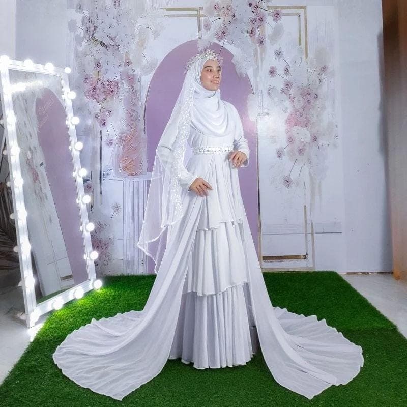 A layered white modest bridal dress with a long detachable train, beaded waistband, and matching veil displayed in a studio setting.