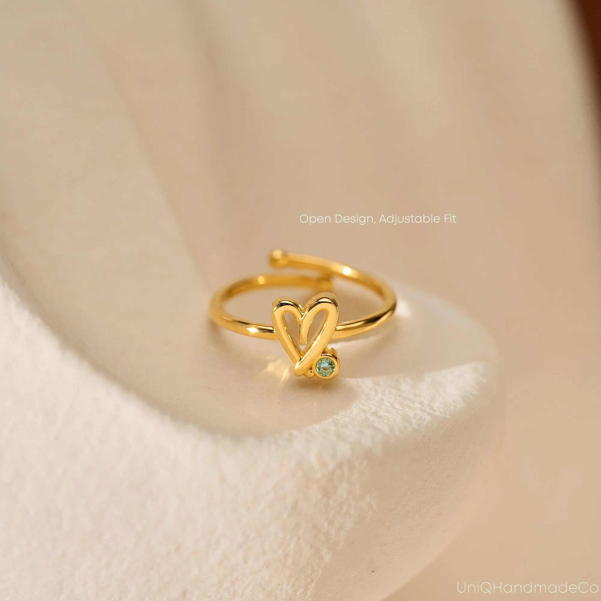 May include: A gold ring featuring a heart-shaped design and a small, round, green gemstone. The ring has an open design and is adjustable. The text "Open Design, Adjustable Fit" is visible.