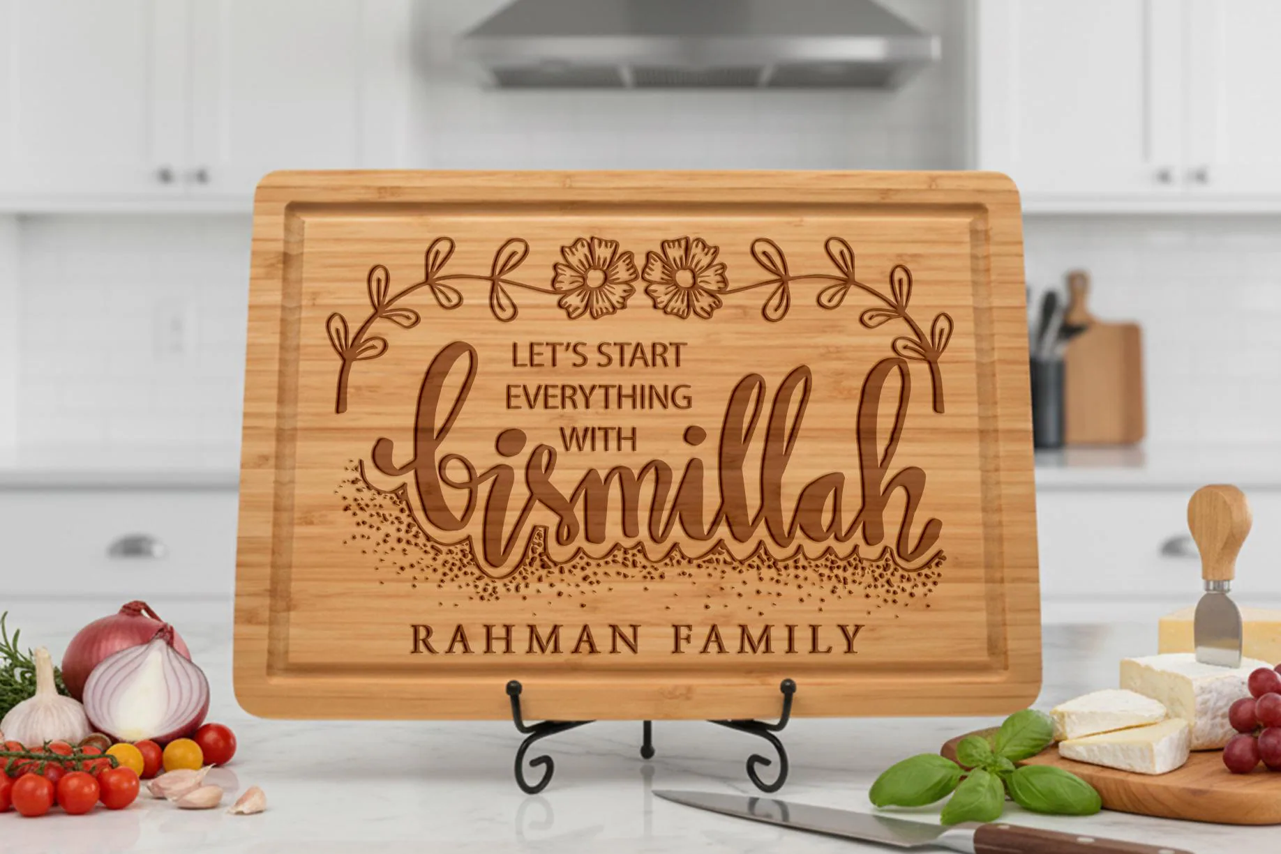 May include: A rectangular bamboo cutting board with a decorative floral design and the words "Let's Start Everything With Bismillah" and "Rahman Family" engraved on it. The board is displayed on a black metal stand.